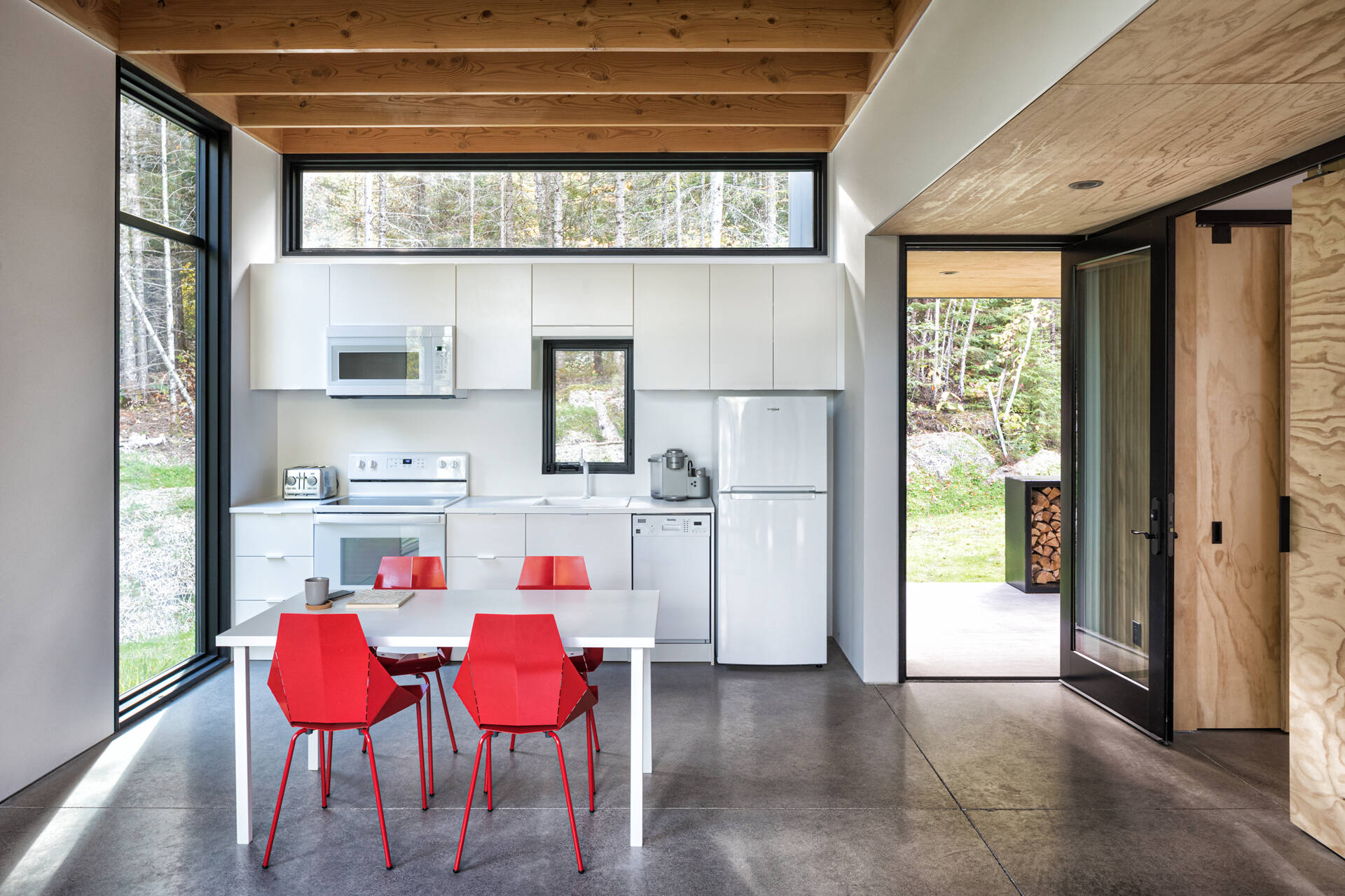 A compact modern kitchen with white cabinetry, black-framed windows, and a simple dining table surrounded by bright red chairs. Polished concrete floors and exposed wood ceiling beams complete the open-plan interior.