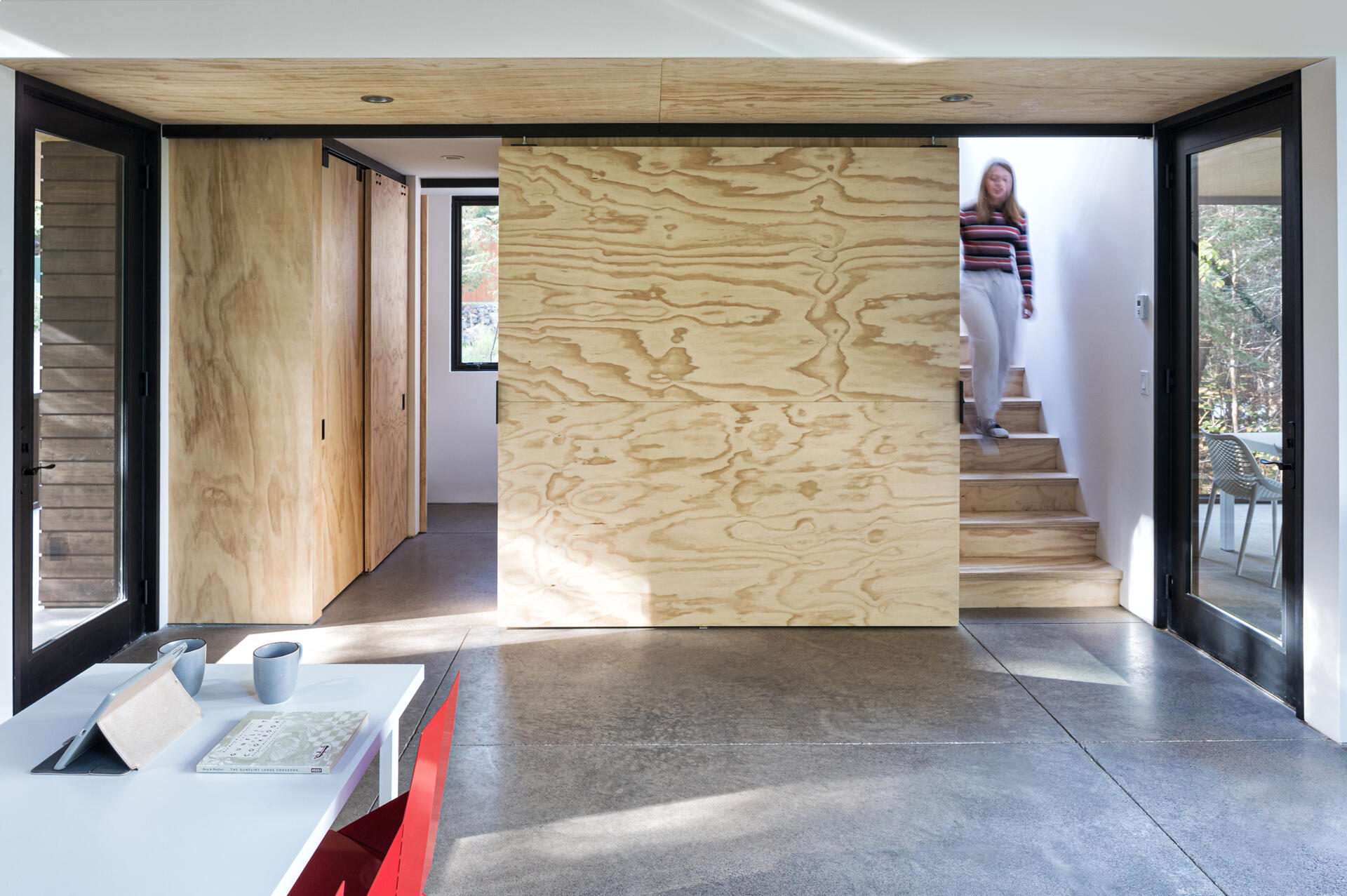 A large sliding plywood door separates the staircase from the main living area of this small modern cabin. Polished concrete flooring, plywood wall panels, and black-framed glass doors define the minimalist interior.