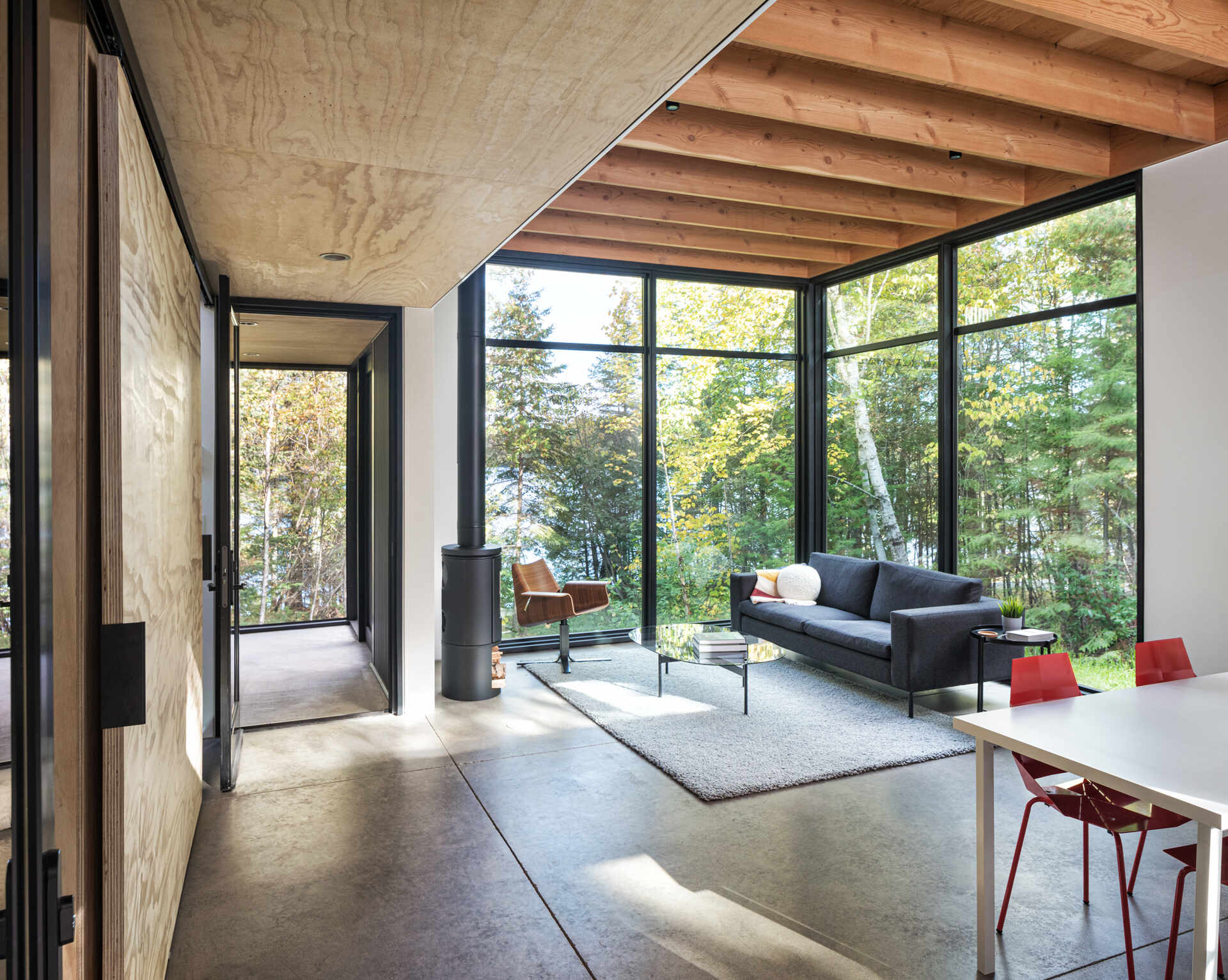 A modern cabin living room with floor-to-ceiling corner windows overlooking the forest, exposed fir ceiling beams, polished concrete floors, and a dark gray sofa. A compact wood-burning stove sits beside the windows.
