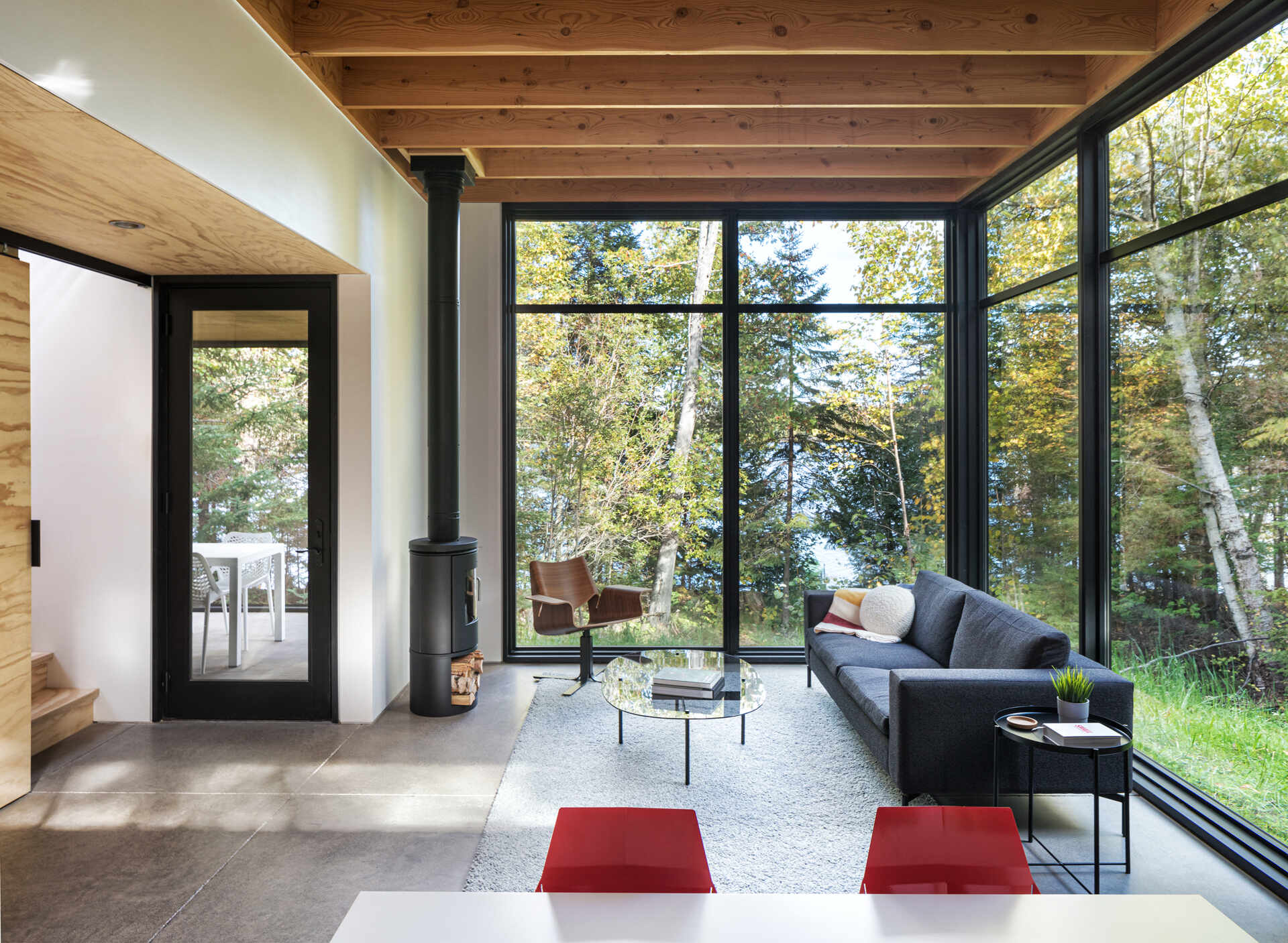 A modern cabin living room with floor-to-ceiling corner windows overlooking the forest, exposed fir ceiling beams, polished concrete floors, and a dark gray sofa. A compact wood-burning stove sits beside the windows.