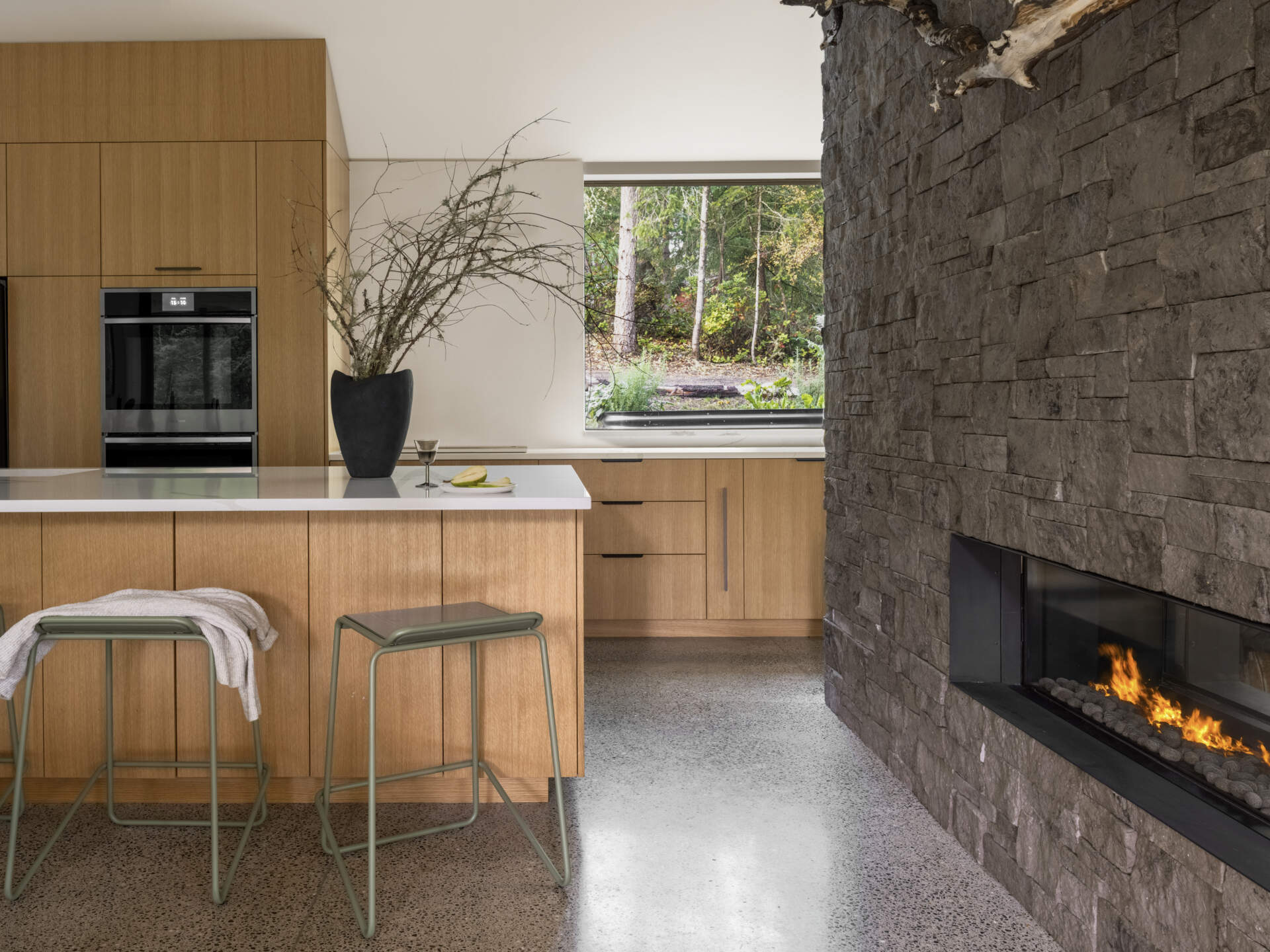 This minimalist modern kitchen pairs natural wood cabinets with white countertops, a central island, polished concrete flooring, and a forest-facing window.