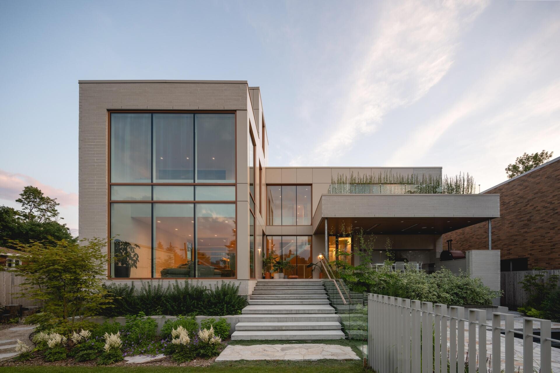Take a look at the backyard of The Lens by MU Architecture, featuring double-height glass walls, a dark-bottomed swimming pool, landscaped gardens, a covered outdoor kitchen, and an upper-level terrace in Montreal.