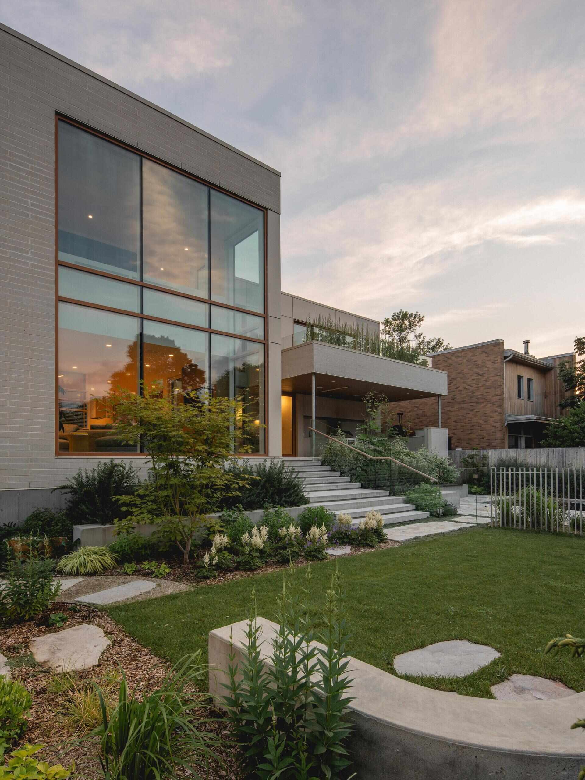 Take a look at the backyard of The Lens by MU Architecture, featuring double-height glass walls, a dark-bottomed swimming pool, landscaped gardens, a covered outdoor kitchen, and an upper-level terrace in Montreal.