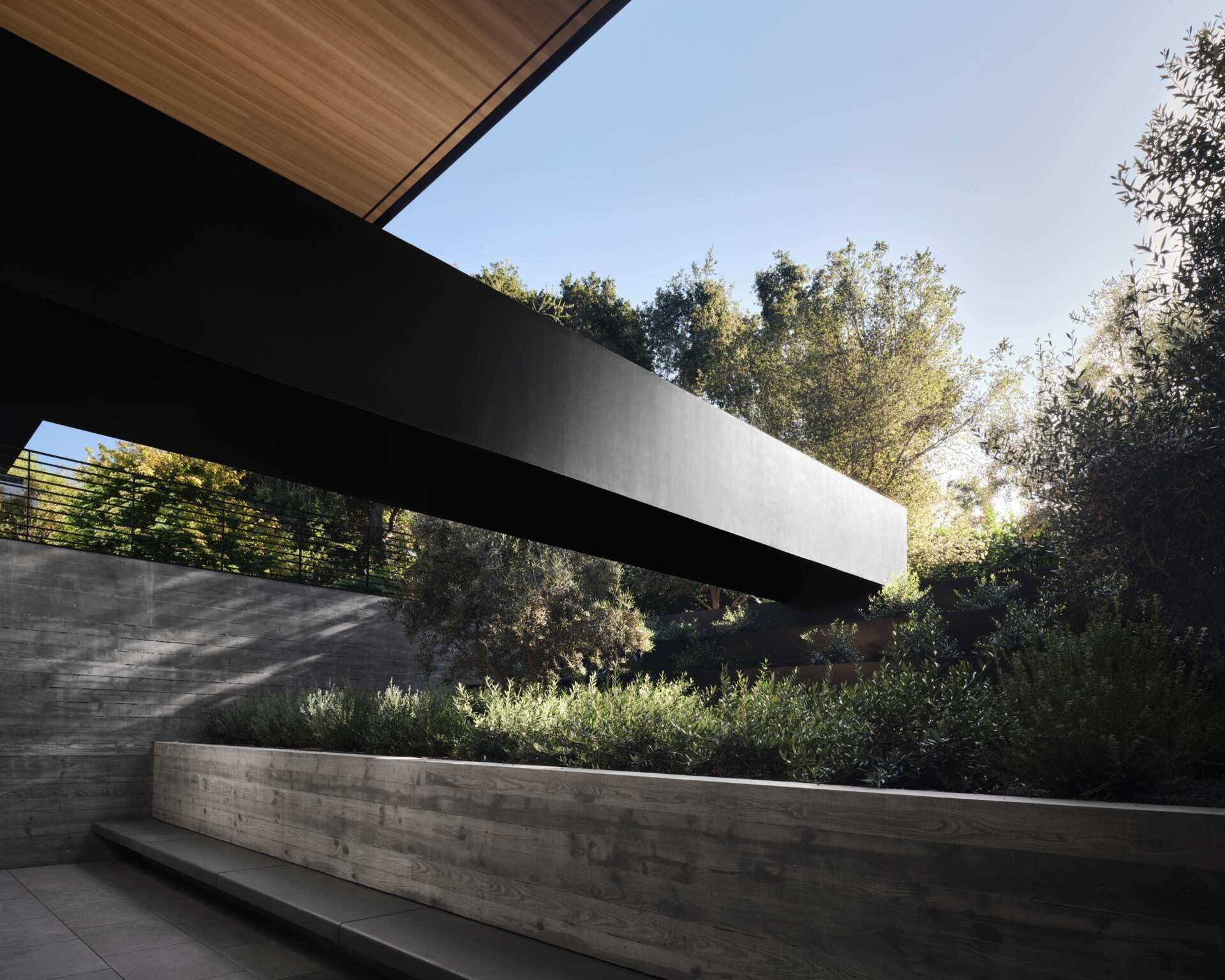 Explore a modern architectural bridge at this Olson Kundig home in California. A bold black steel structure spans a planted garden with board-formed concrete walls, olive trees, built-in seating, and contemporary landscape design.