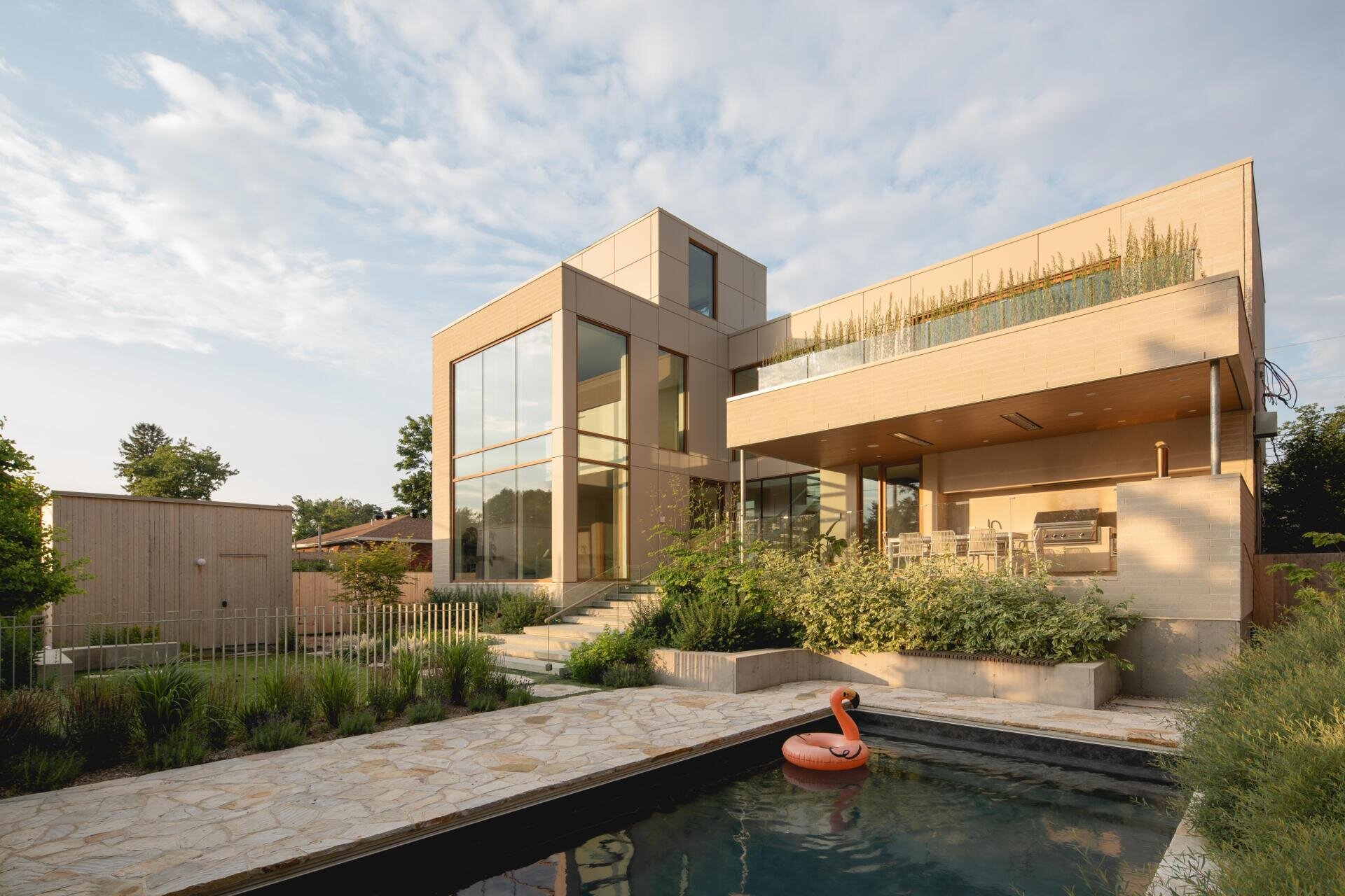 Take a look at the backyard of The Lens by MU Architecture, featuring double-height glass walls, a dark-bottomed swimming pool, landscaped gardens, a covered outdoor kitchen, and an upper-level terrace in Montreal.