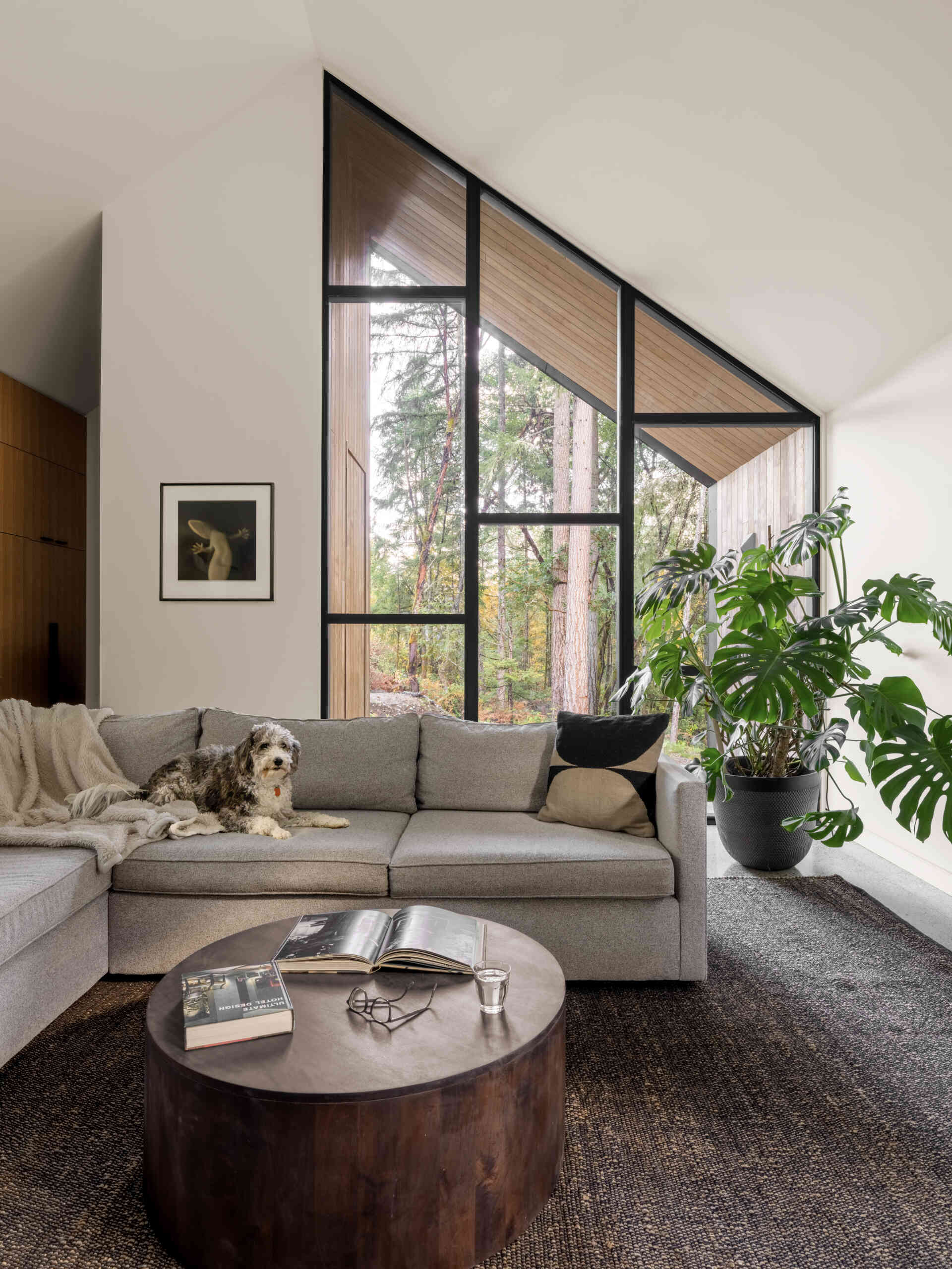 Step inside a modern forest living room with a vaulted ceiling, grey sectional sofa, sculptural wooden furniture, a large stone fireplace, and dramatic floor-to-ceiling windows.