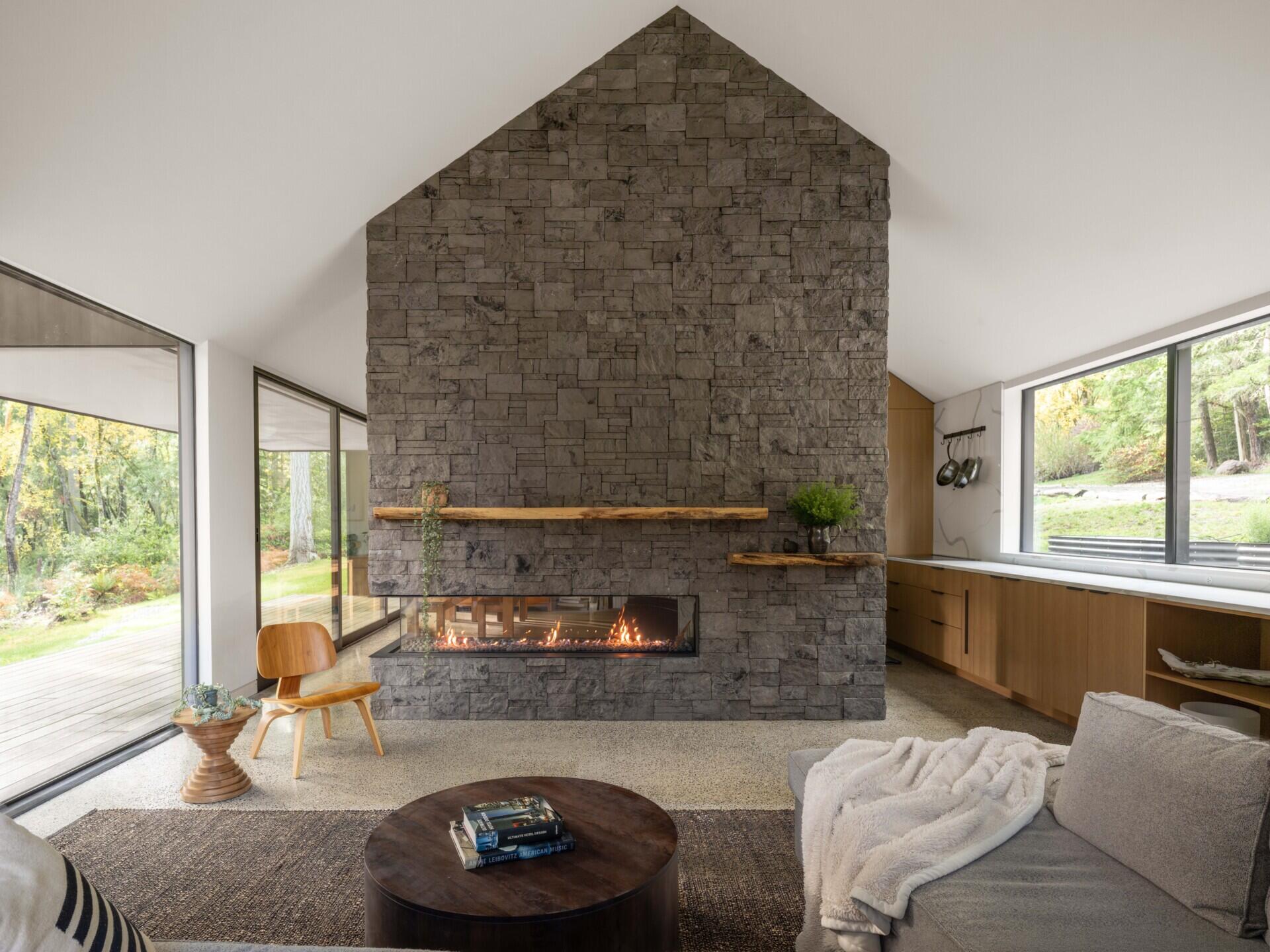 Step inside a modern forest living room with a vaulted ceiling, grey sectional sofa, sculptural wooden furniture, a large stone fireplace, and dramatic floor-to-ceiling windows.