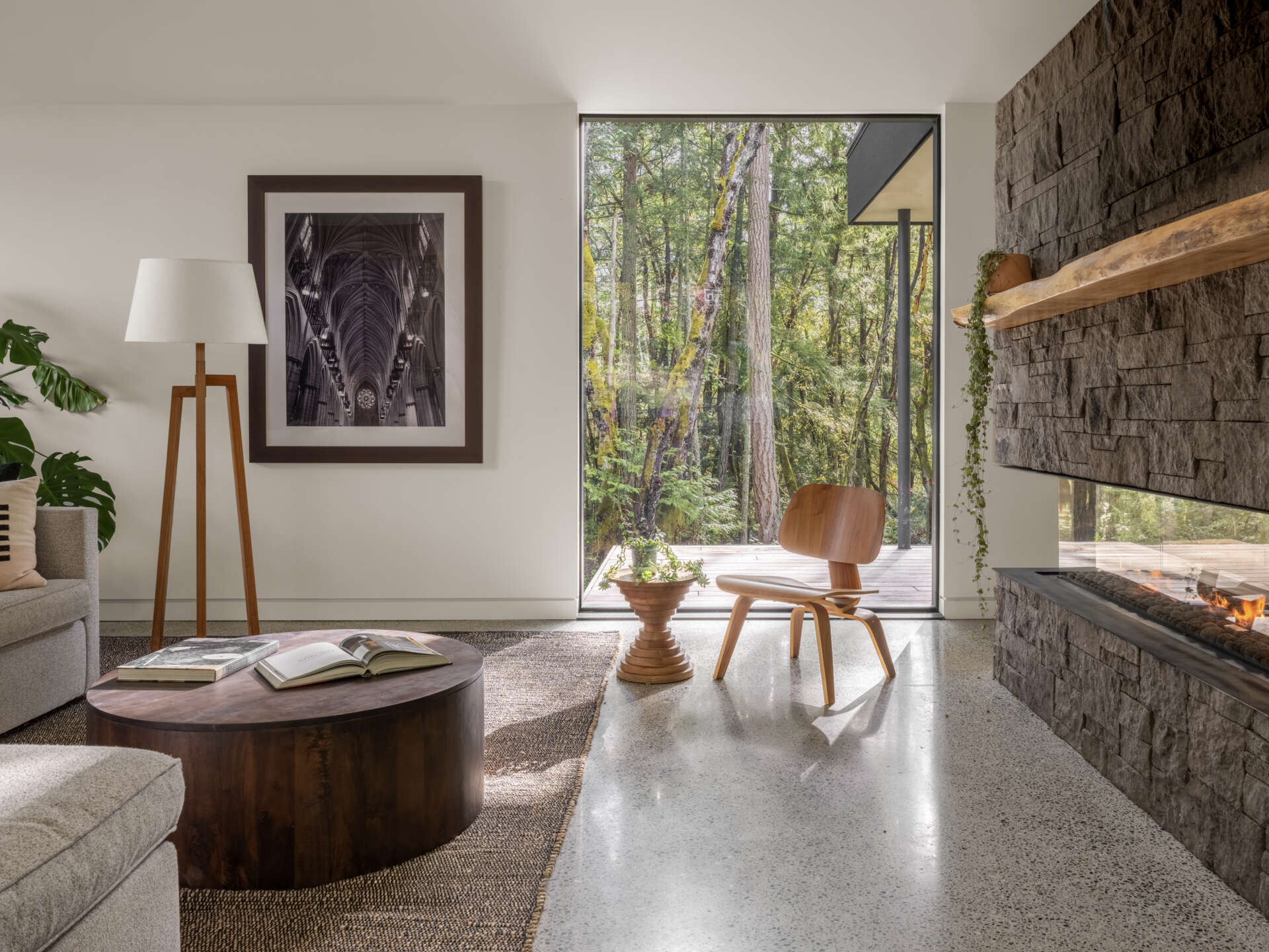 Step inside a modern forest living room with a vaulted ceiling, grey sectional sofa, sculptural wooden furniture, a large stone fireplace, and dramatic floor-to-ceiling windows.