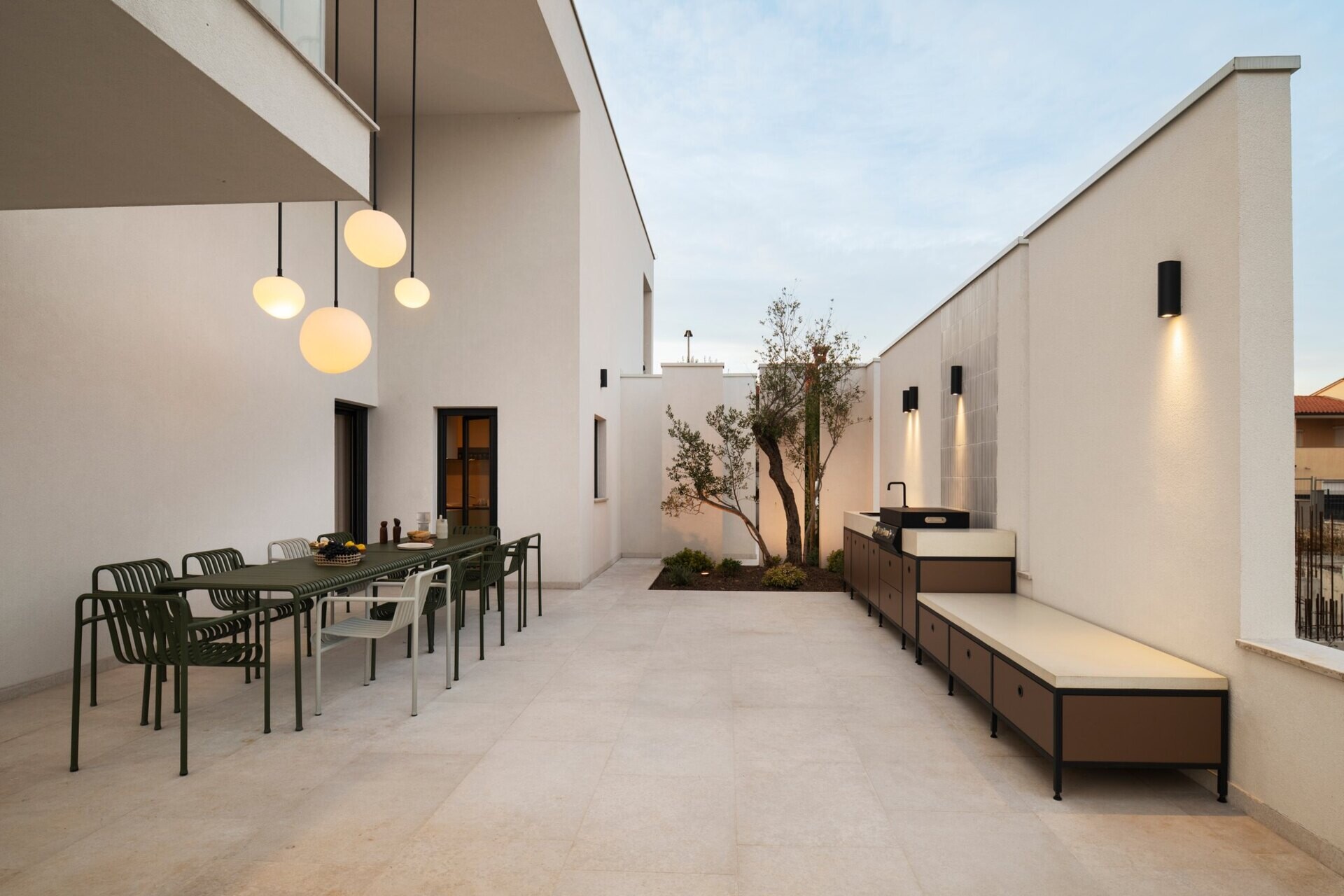 Take a look at the outdoor patios of Arc House in Croatia, featuring a Mediterranean dining terrace with an outdoor kitchen, modern green furniture, globe pendant lights and a covered lounge with white seating and garden views.