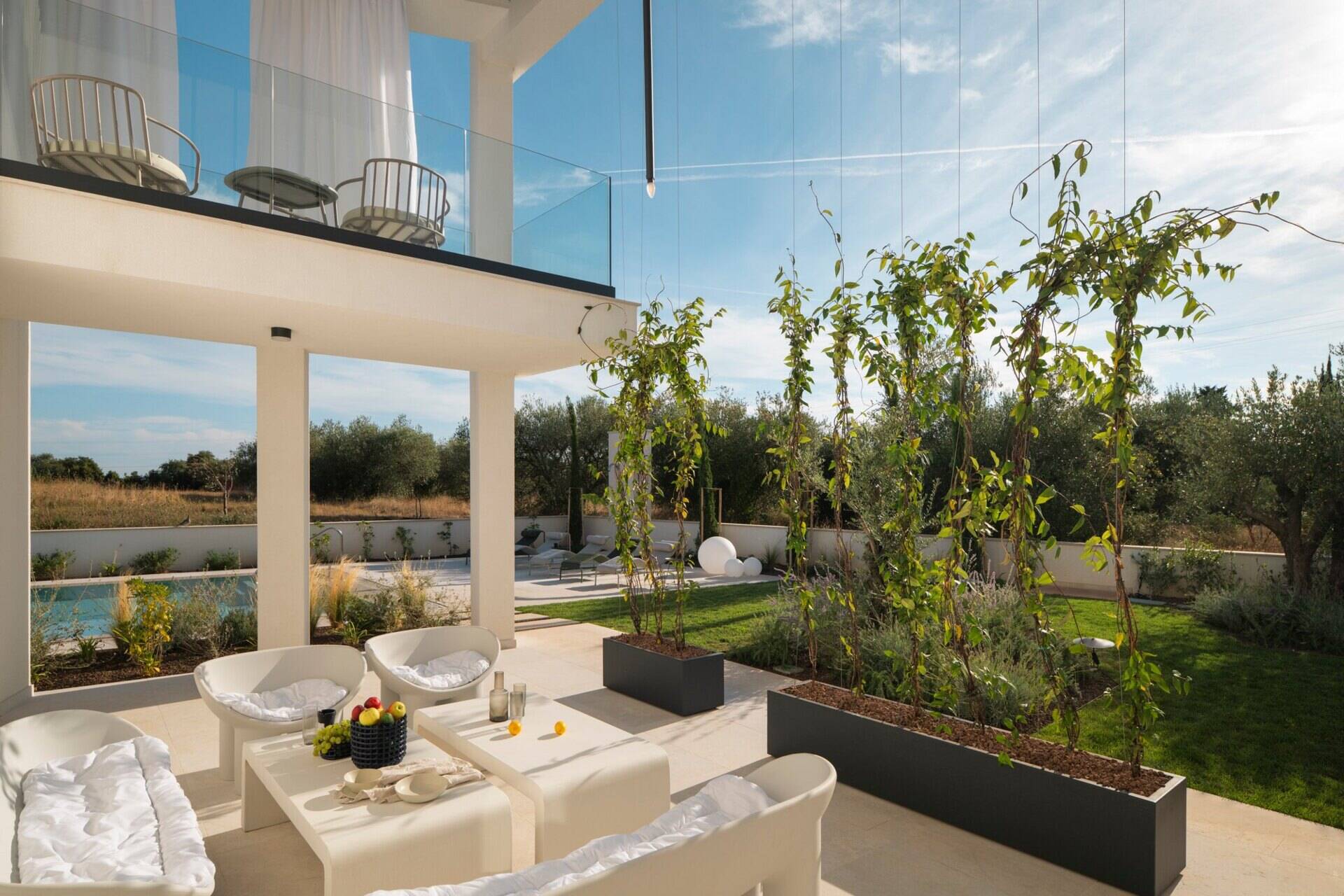 Take a look at the outdoor patios of Arc House in Croatia, featuring a Mediterranean dining terrace with an outdoor kitchen, modern green furniture, globe pendant lights and a covered lounge with white seating and garden views.