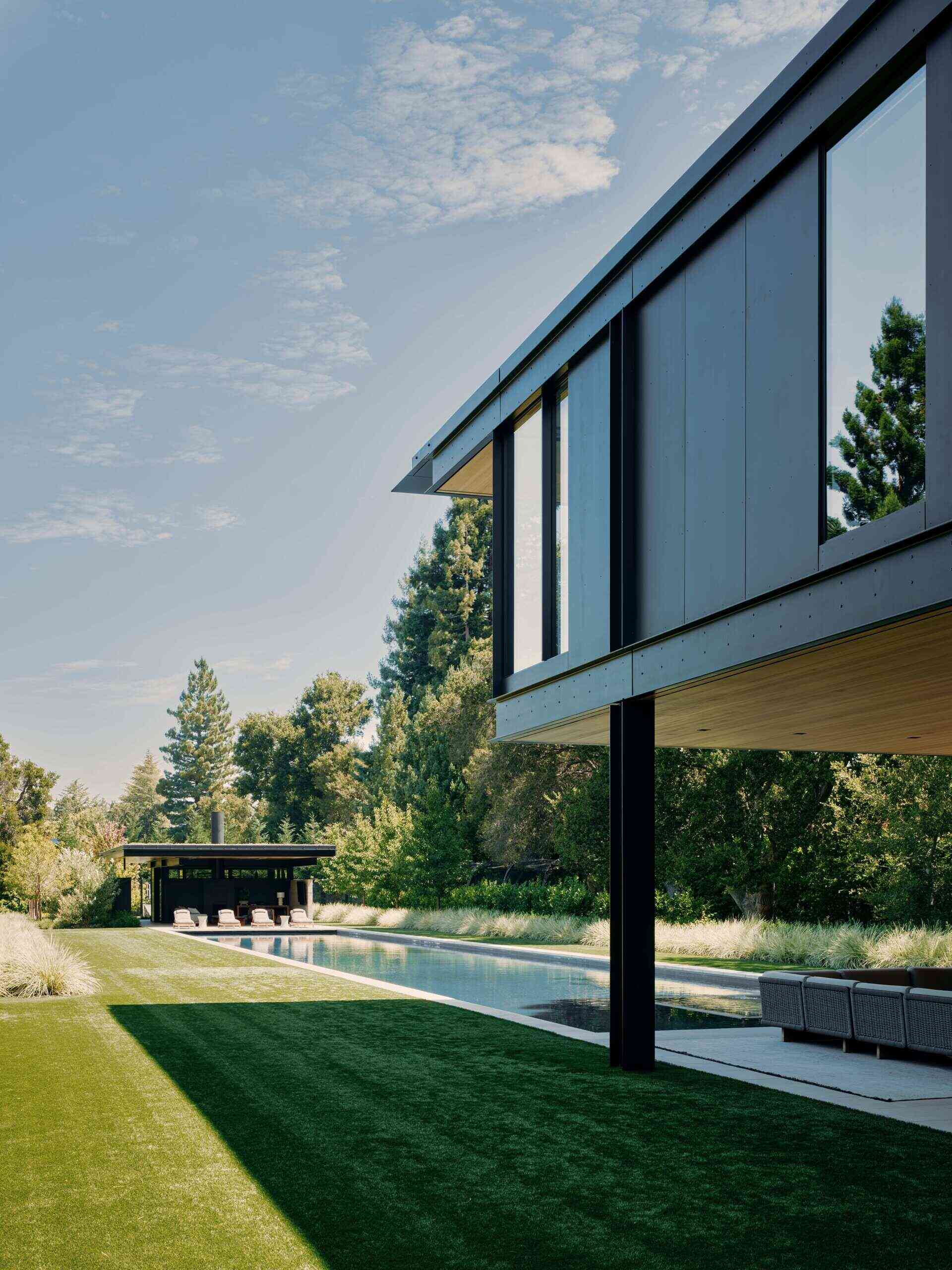 Explore a modern swimming pool and pool house at this California home by Olson Kundig. A long rectangular pool, contemporary sun loungers, landscaped lawn, mature trees, and a pavilion with a green roof create an impressive outdoor leisure area.