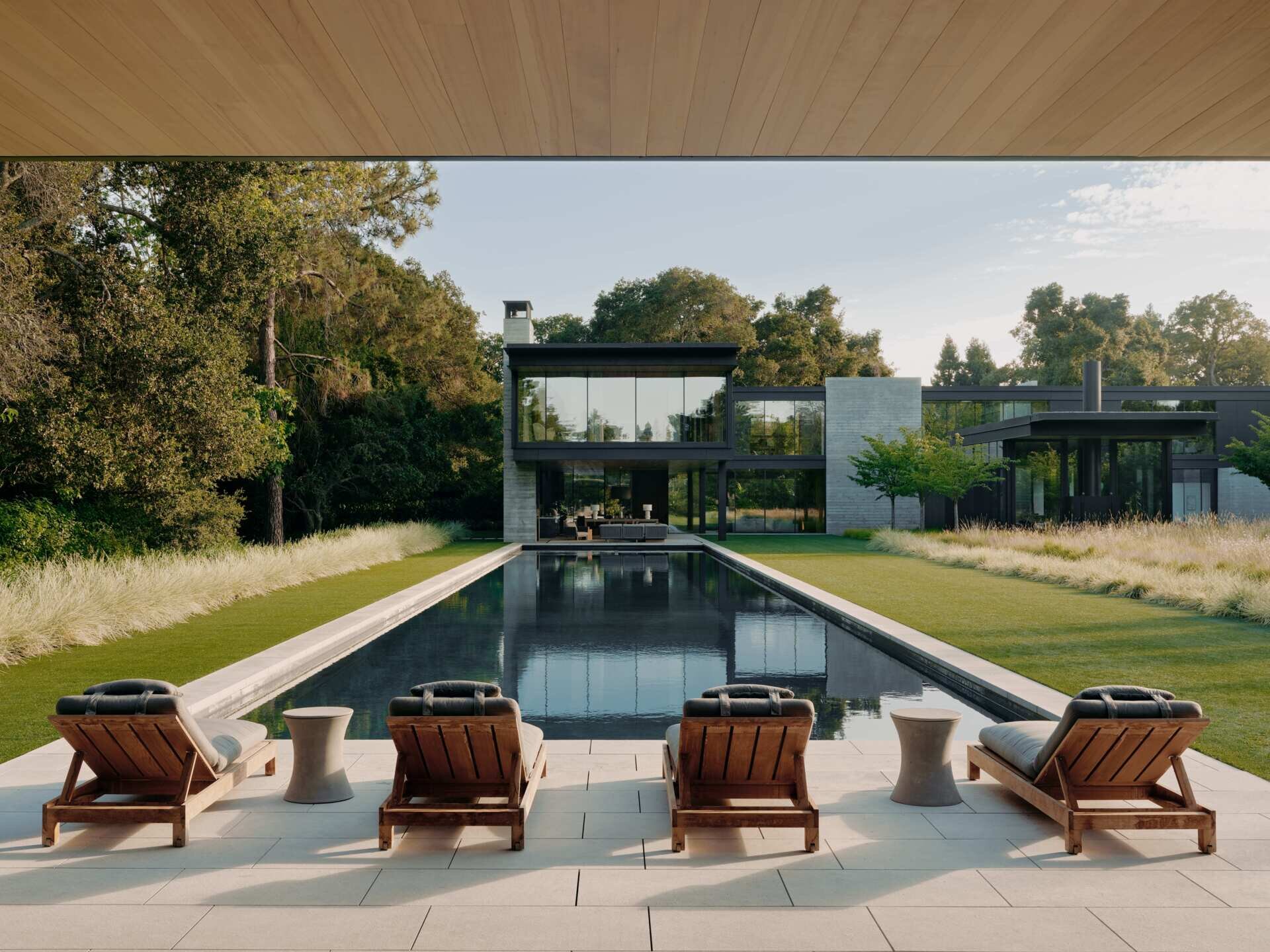 Explore a modern swimming pool and pool house at this California home by Olson Kundig. A long rectangular pool, contemporary sun loungers, landscaped lawn, mature trees, and a pavilion with a green roof create an impressive outdoor leisure area.
