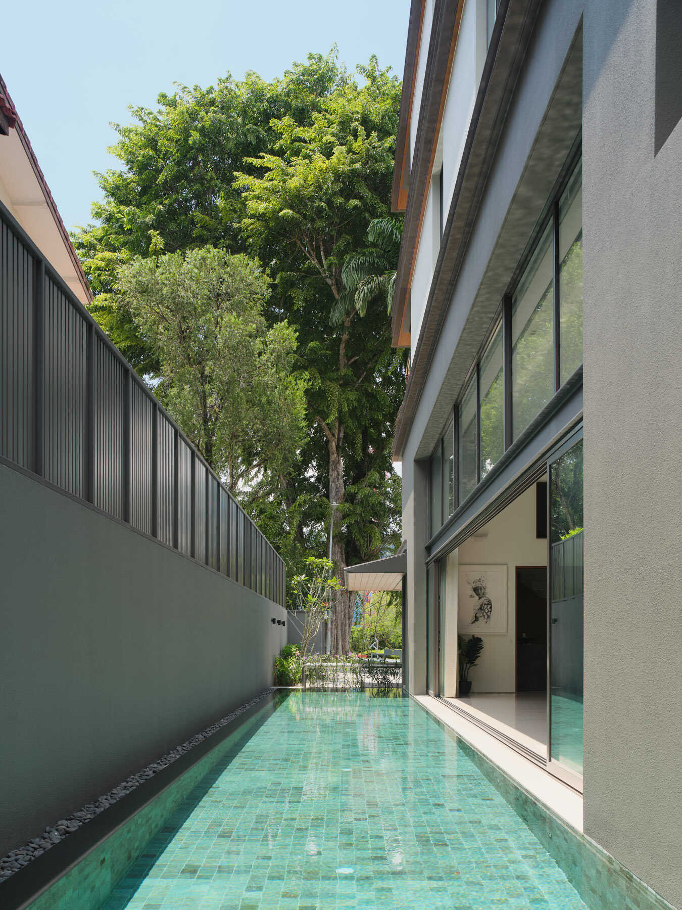 See the long, narrow swimming pool at JS-House in Singapore, with turquoise tiles, floor-to-ceiling sliding glass doors, privacy screening, and lush tropical landscaping.