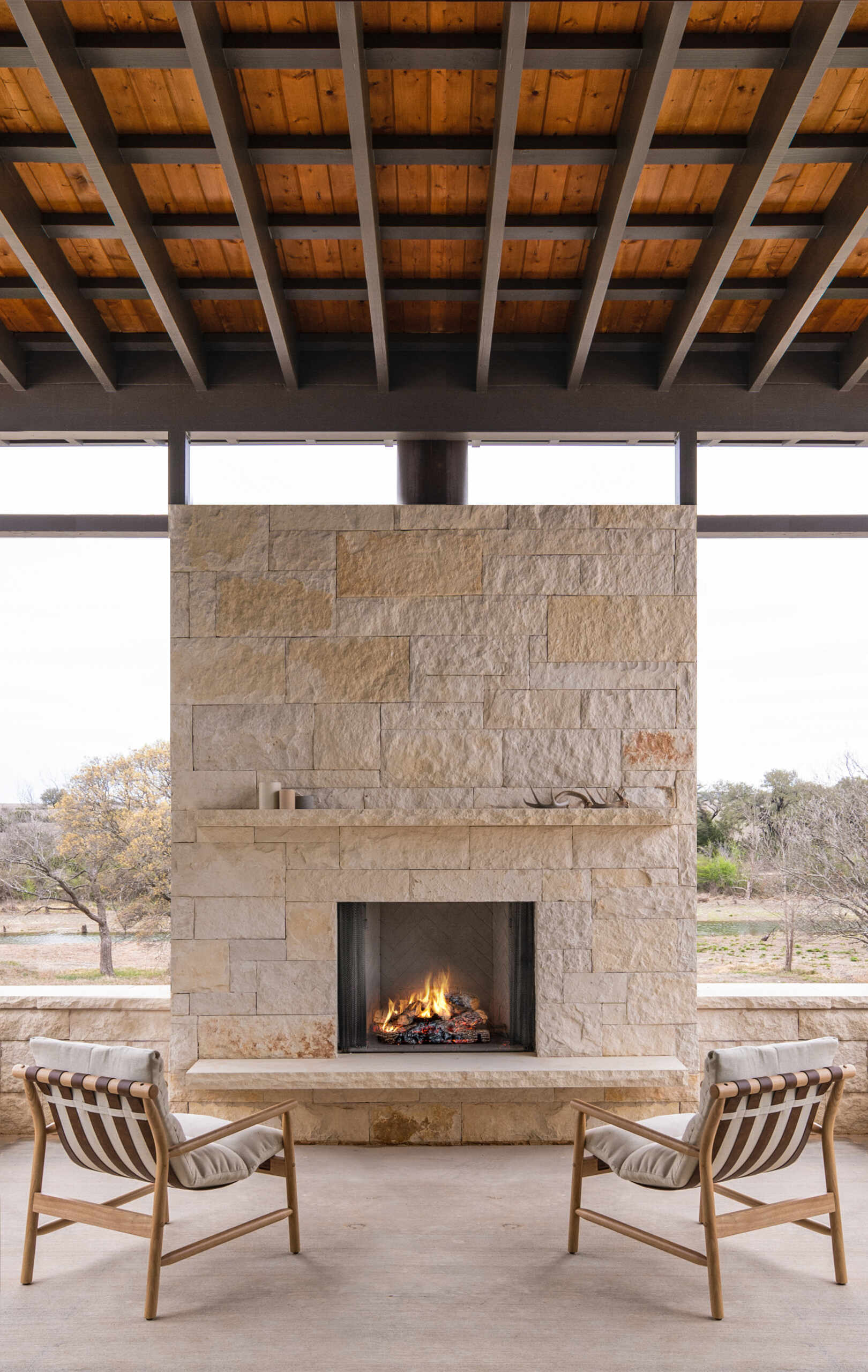 A covered outdoor sitting area features a limestone fireplace, exposed ceiling structure, and views of the Texas landscape.