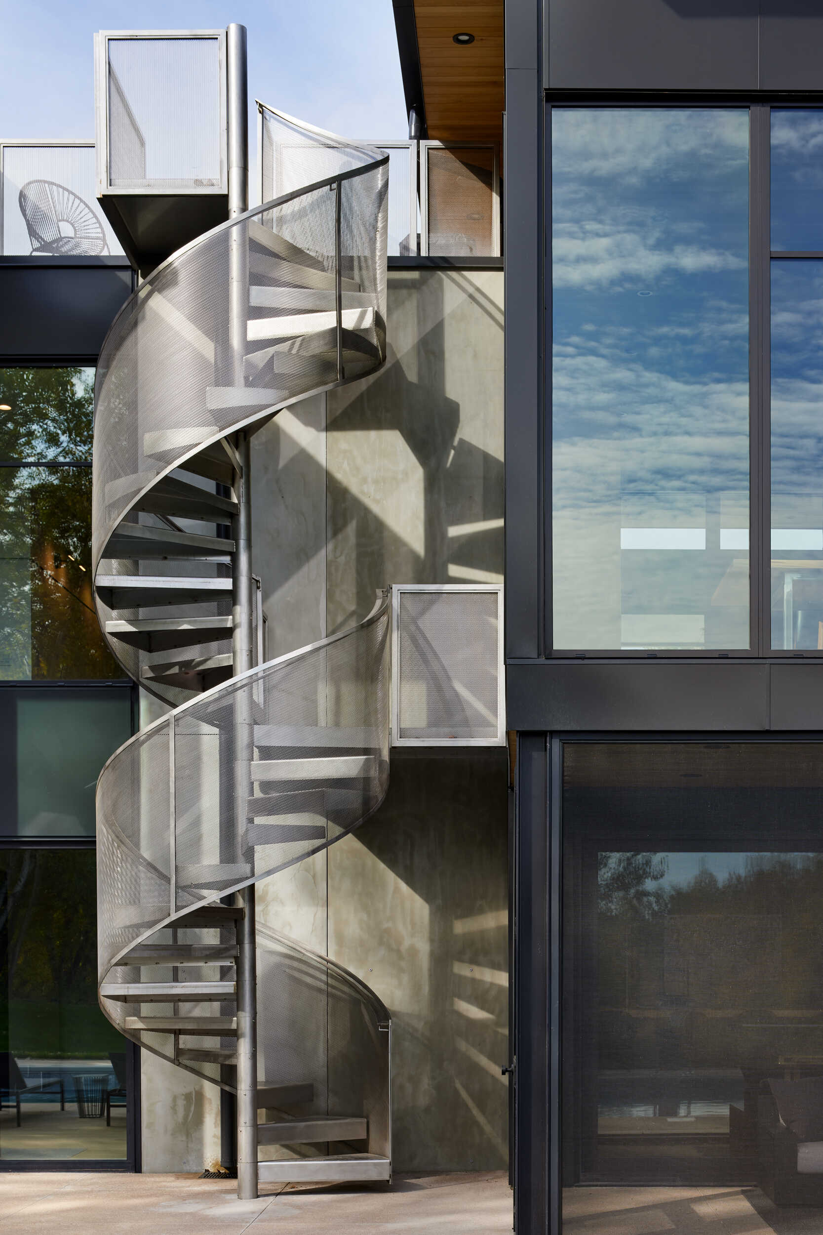 A close-up of a stainless steel spiral staircase with perforated metal railings and open steps. The exterior staircase leads to a rooftop deck between two sections of a modern home.