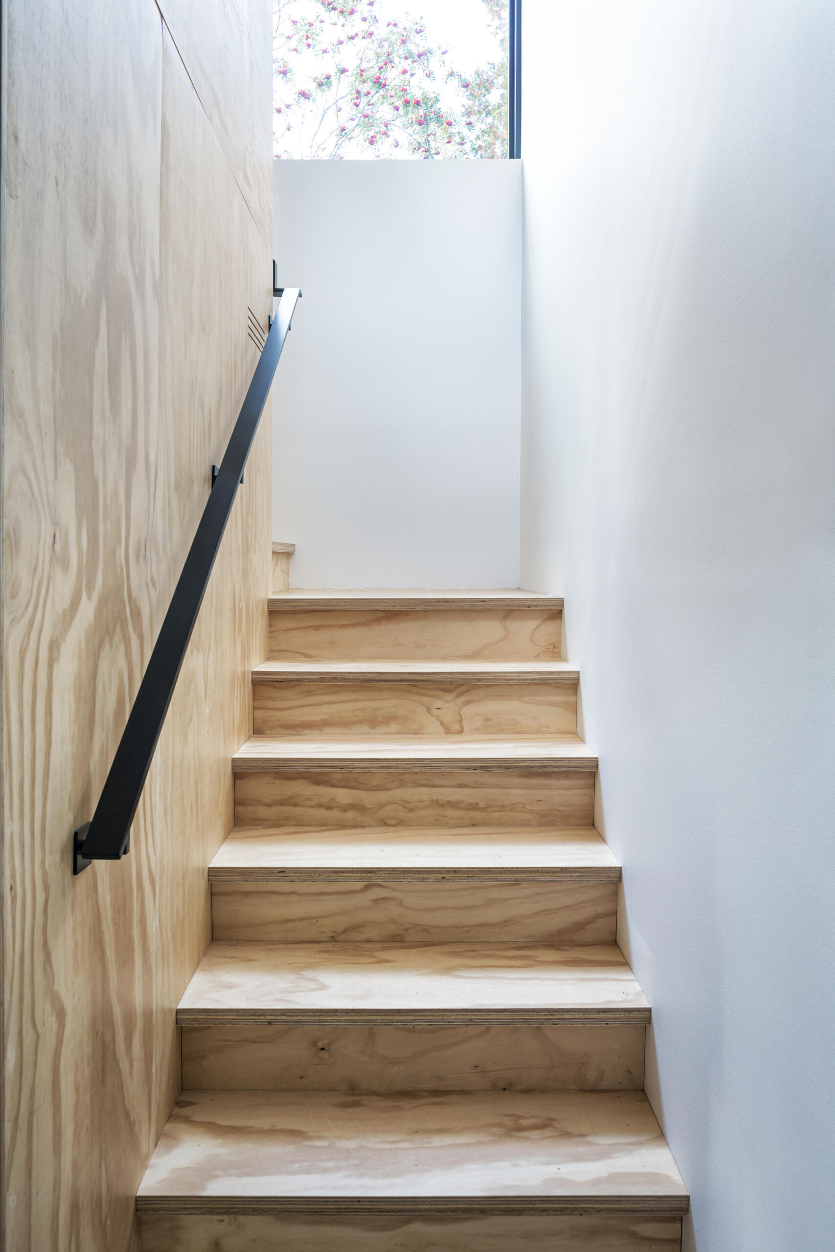 A narrow plywood staircase with exposed wood grain, a black metal handrail, and white walls. A window above the landing brings natural light into the stairwell.