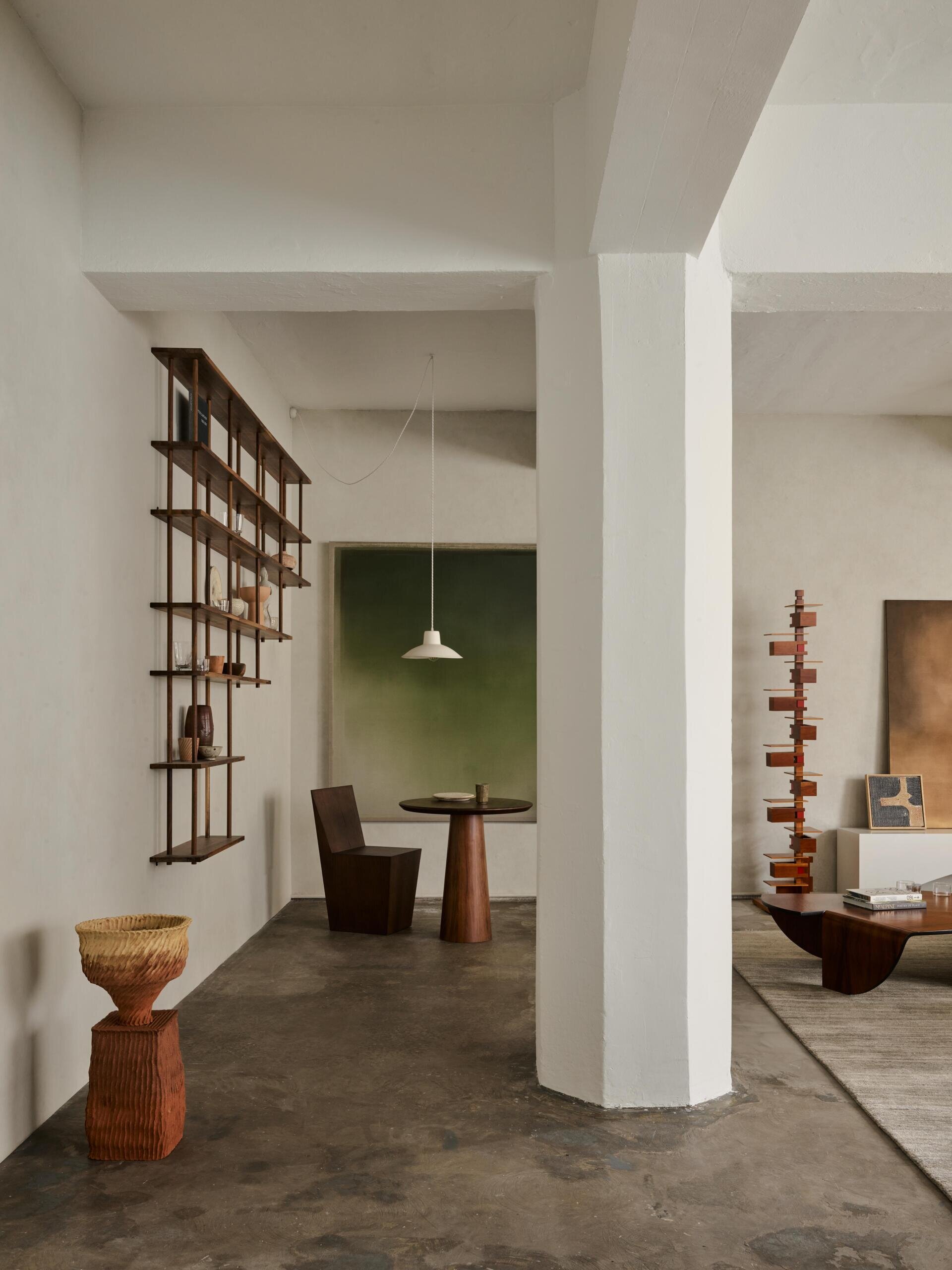 An industrial-style apartment interior with exposed concrete columns and ceiling beams painted white, open wood display shelving, sculptural furniture, and a polished concrete floor. The open layout connects a small seating area with the contemporary living room.