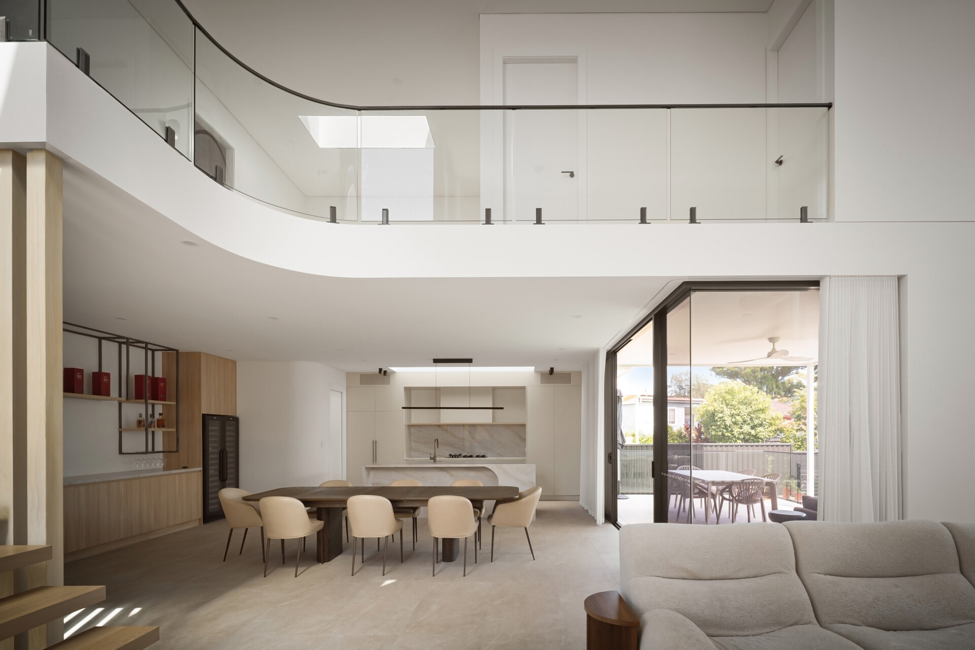 A modern open-plan interior with a double-height living area, curved upstairs balcony, and glass railing overlooking the dining room and kitchen. Pale flooring, white walls, wood cabinetry, and large sliding glass doors create a bright contemporary space.