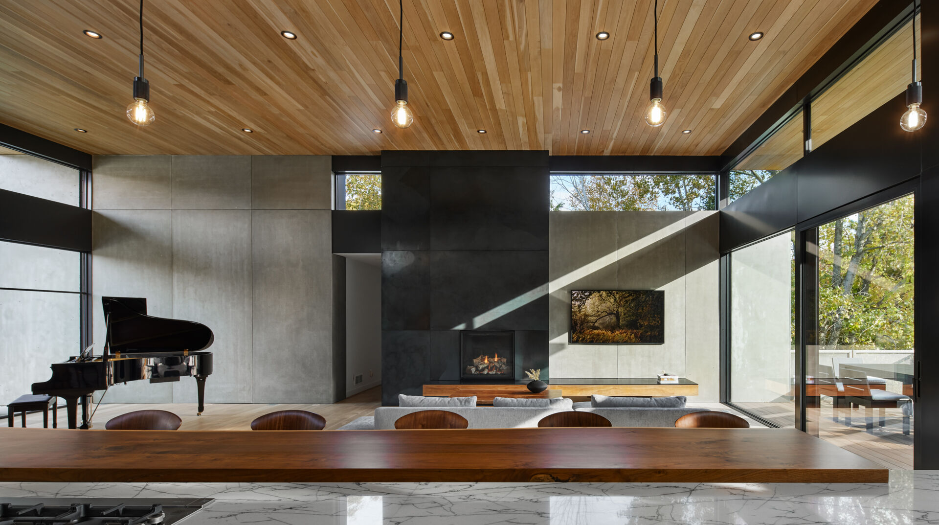 A modern open-plan interior with a black grand piano, a tall fireplace wall clad in contrasting concrete panels, and a wood ceiling. Large windows and glass doors bring daylight into the living area.