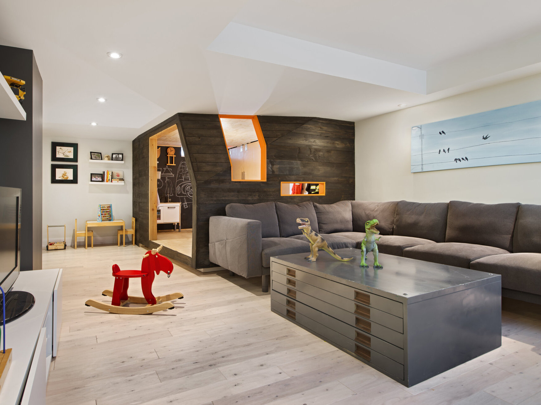 A creative children's playhouse built inside an Ottawa basement features dark reclaimed wood, bright orange windows, plywood interiors, and built-in toy storage. Designed by 25:8 Architecture + Urban Design, the miniature house creates a fun play area beside the family living room.
