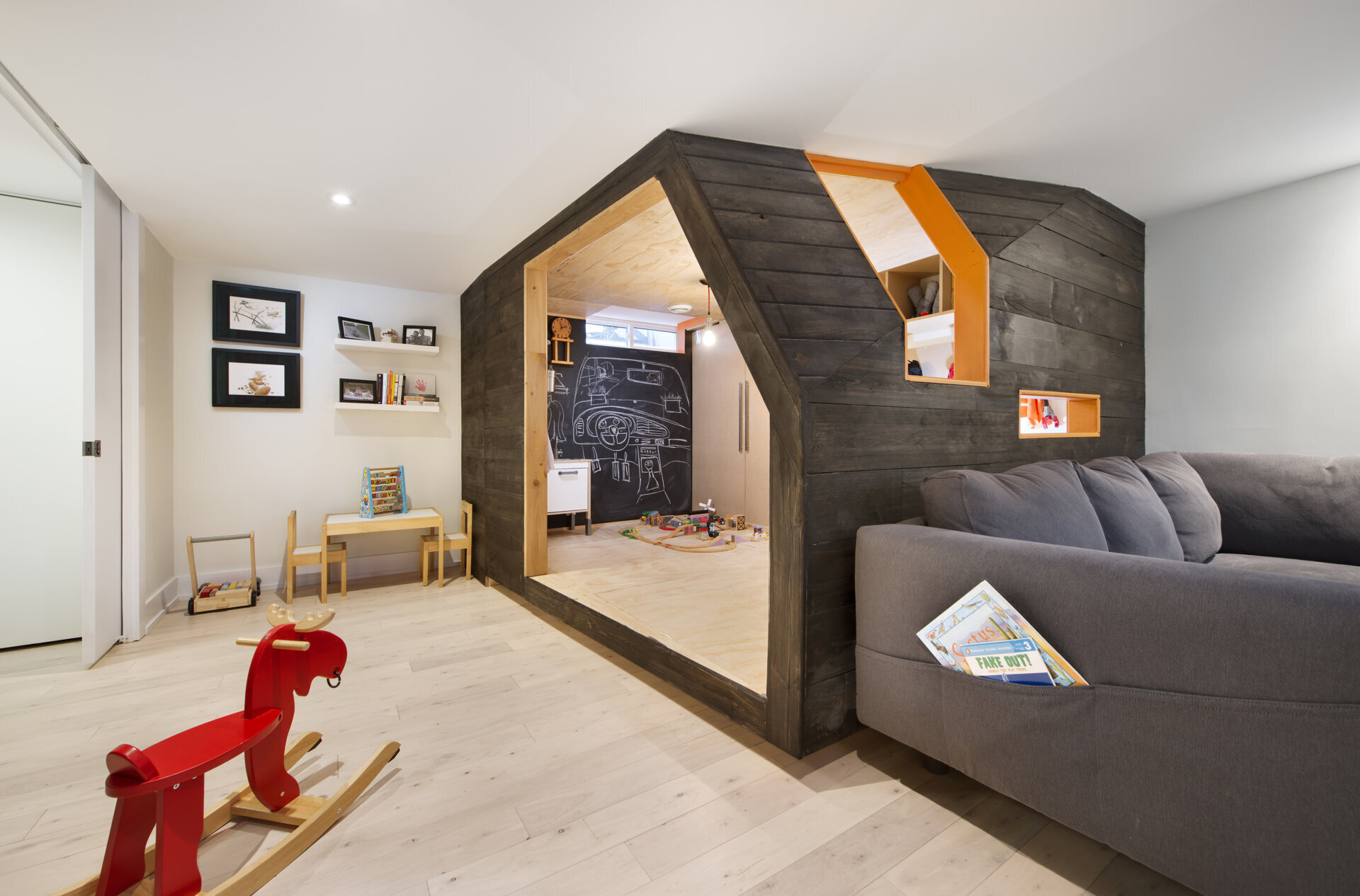A creative children's playhouse built inside an Ottawa basement features dark reclaimed wood, bright orange windows, plywood interiors, and built-in toy storage. Designed by 25:8 Architecture + Urban Design, the miniature house creates a fun play area beside the family living room.