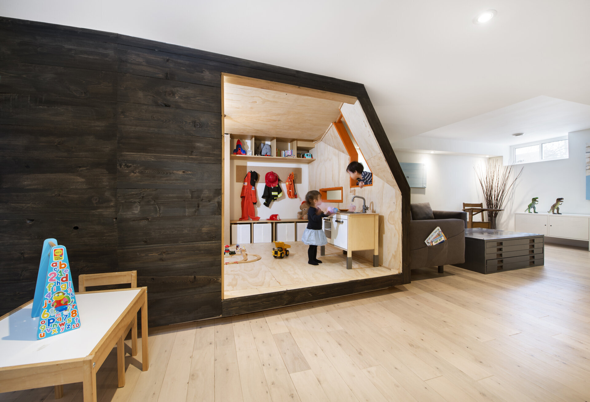 A creative children's playhouse built inside an Ottawa basement features dark reclaimed wood, bright orange windows, plywood interiors, and built-in toy storage. Designed by 25:8 Architecture + Urban Design, the miniature house creates a fun play area beside the family living room.