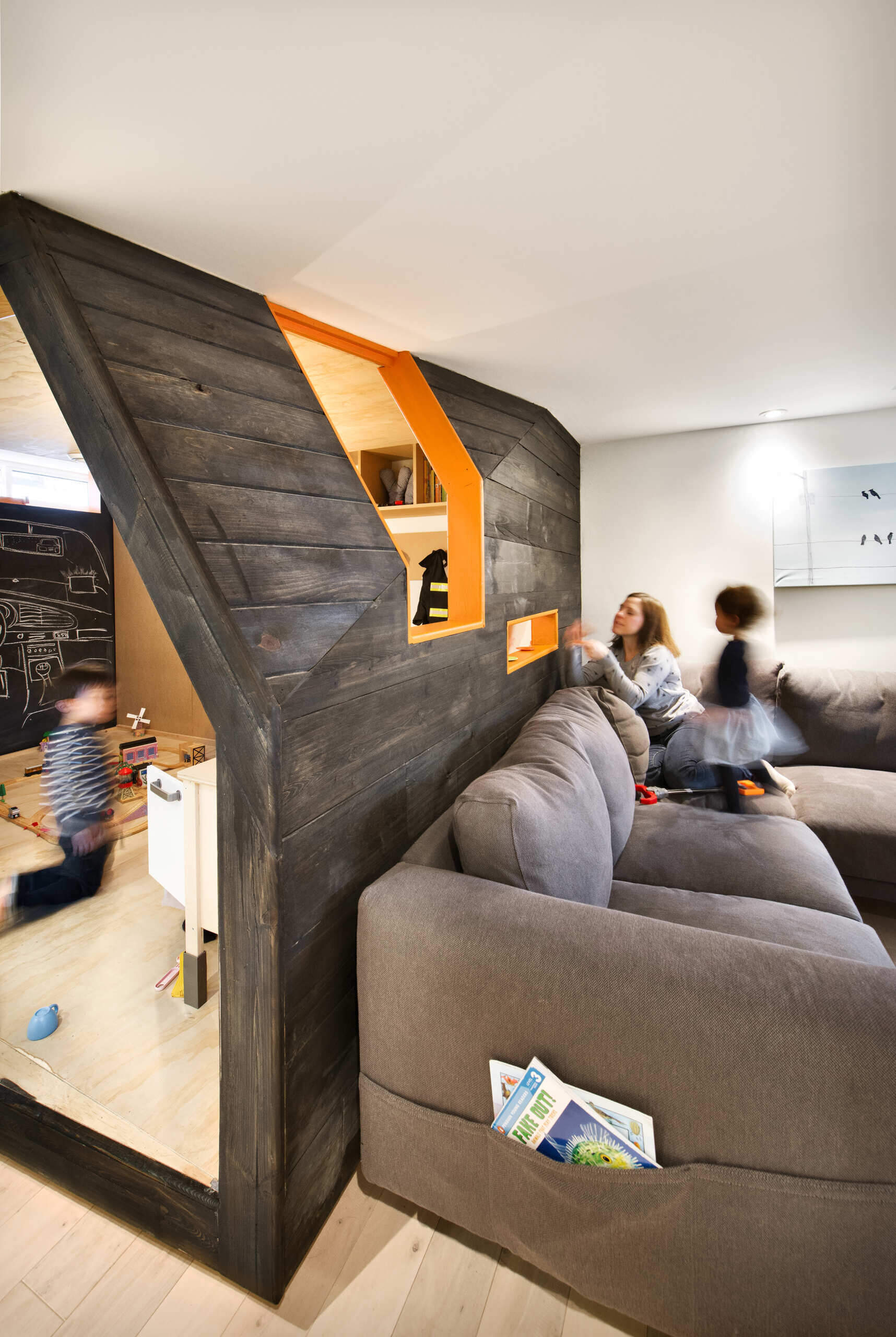A creative children's playhouse built inside an Ottawa basement features dark reclaimed wood, bright orange windows, plywood interiors, and built-in toy storage. Designed by 25:8 Architecture + Urban Design, the miniature house creates a fun play area beside the family living room.
