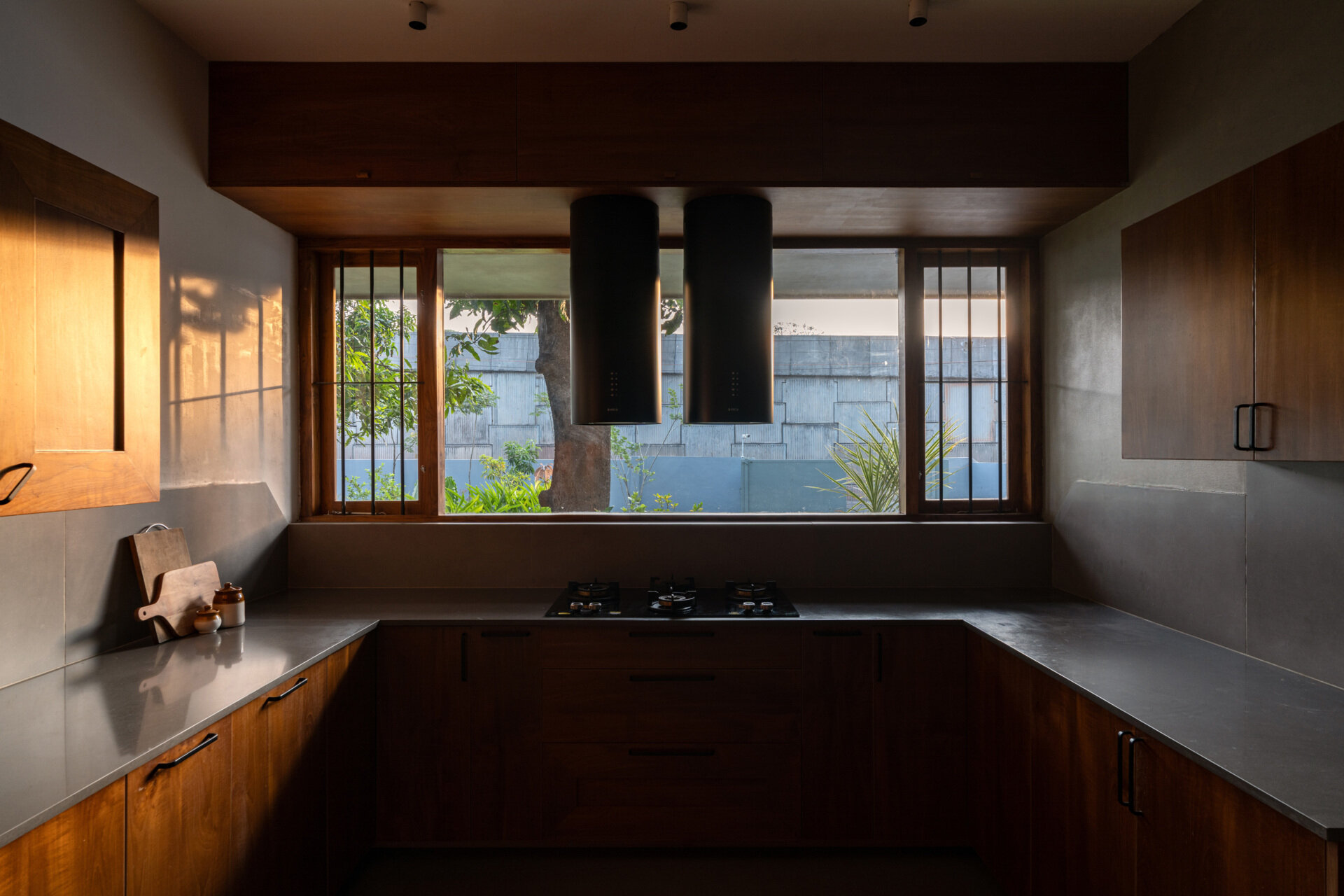 This contemporary U-shaped kitchen combines wood cabinets, quartz countertops, black range hoods, and a large garden-facing window that welcomes morning sunlight.