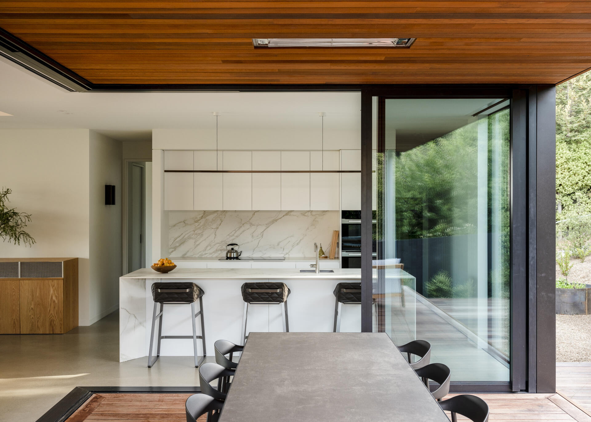 Modern white kitchen with marble countertops, a large island, and black bar stools opening onto a covered outdoor dining area. Sliding glass doors connect the kitchen to the wood deck and surrounding greenery.