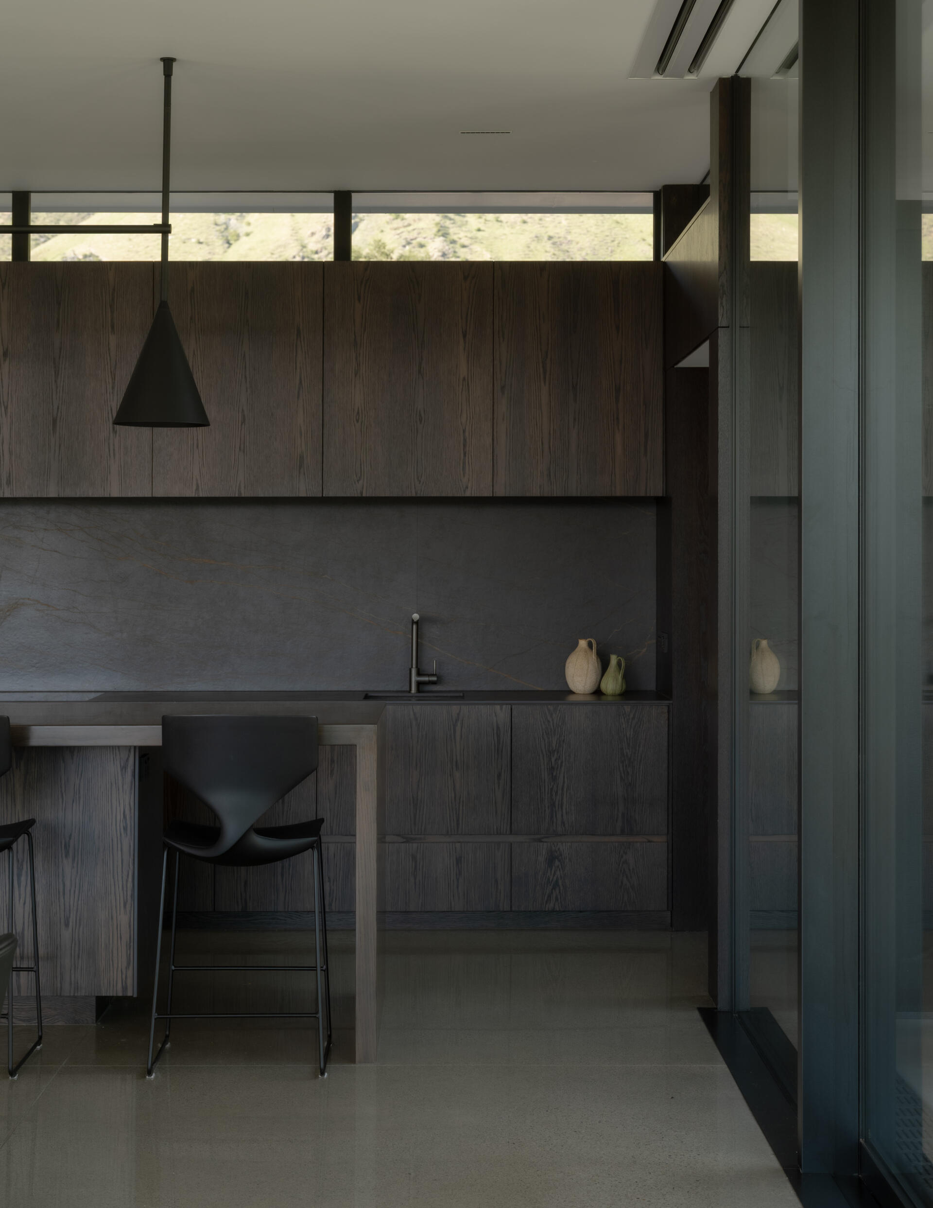 A contemporary dark oak kitchen with black marble countertops and backsplash, minimalist cabinetry, a long breakfast bar, black pendant lights, and polished concrete floors.