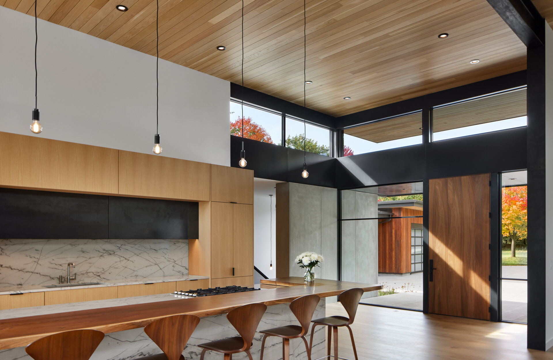 A modern kitchen with light wood cabinetry, a marble-look island, and a long wood breakfast bar lined with sculptural stools. High windows, exposed steel beams, and a wood ceiling define the open interior.