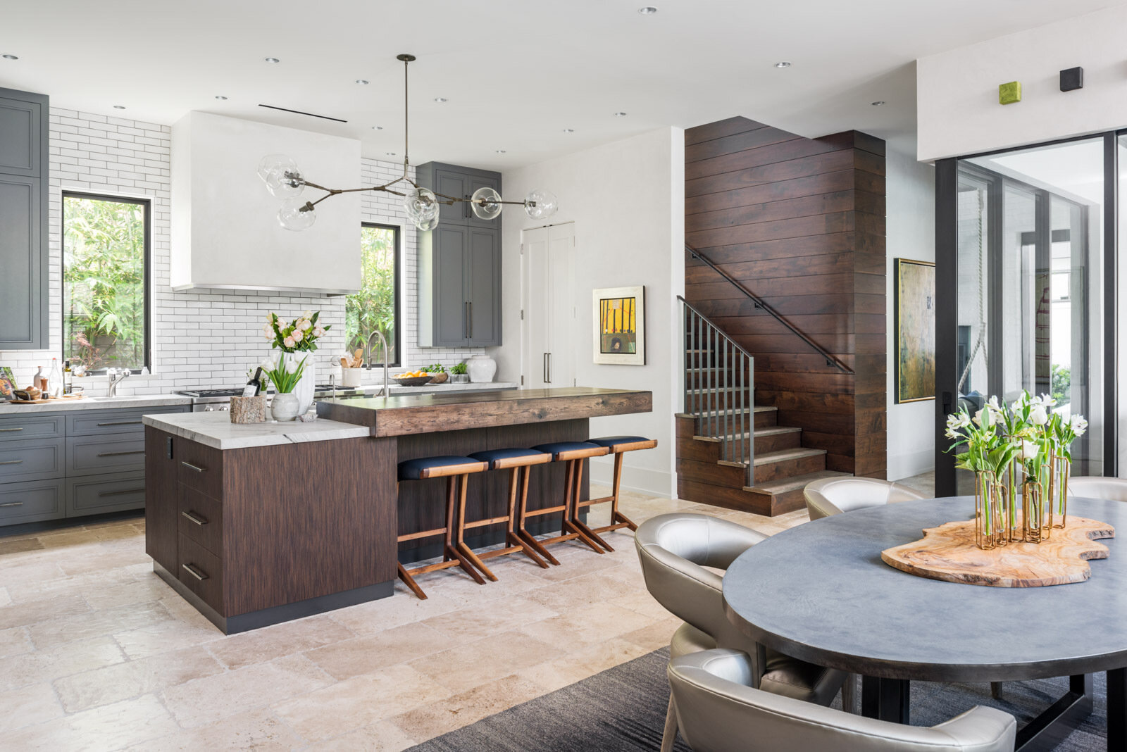 A contemporary kitchen with gray cabinets, white subway tile, stone countertops, a dark wood island, rustic wood breakfast bar, and modern glass globe pendant lighting.