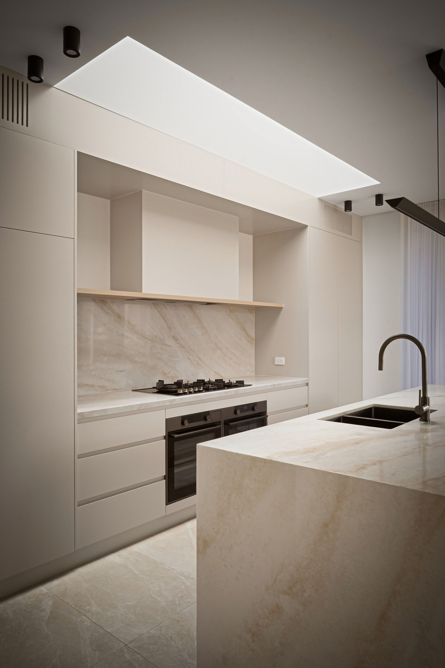 A modern minimalist kitchen featuring a large rectangular skylight above white cabinetry and a natural stone backsplash. The stone island includes an integrated sink and tall metal faucet, with dark appliances and a slim pendant light providing contrast.