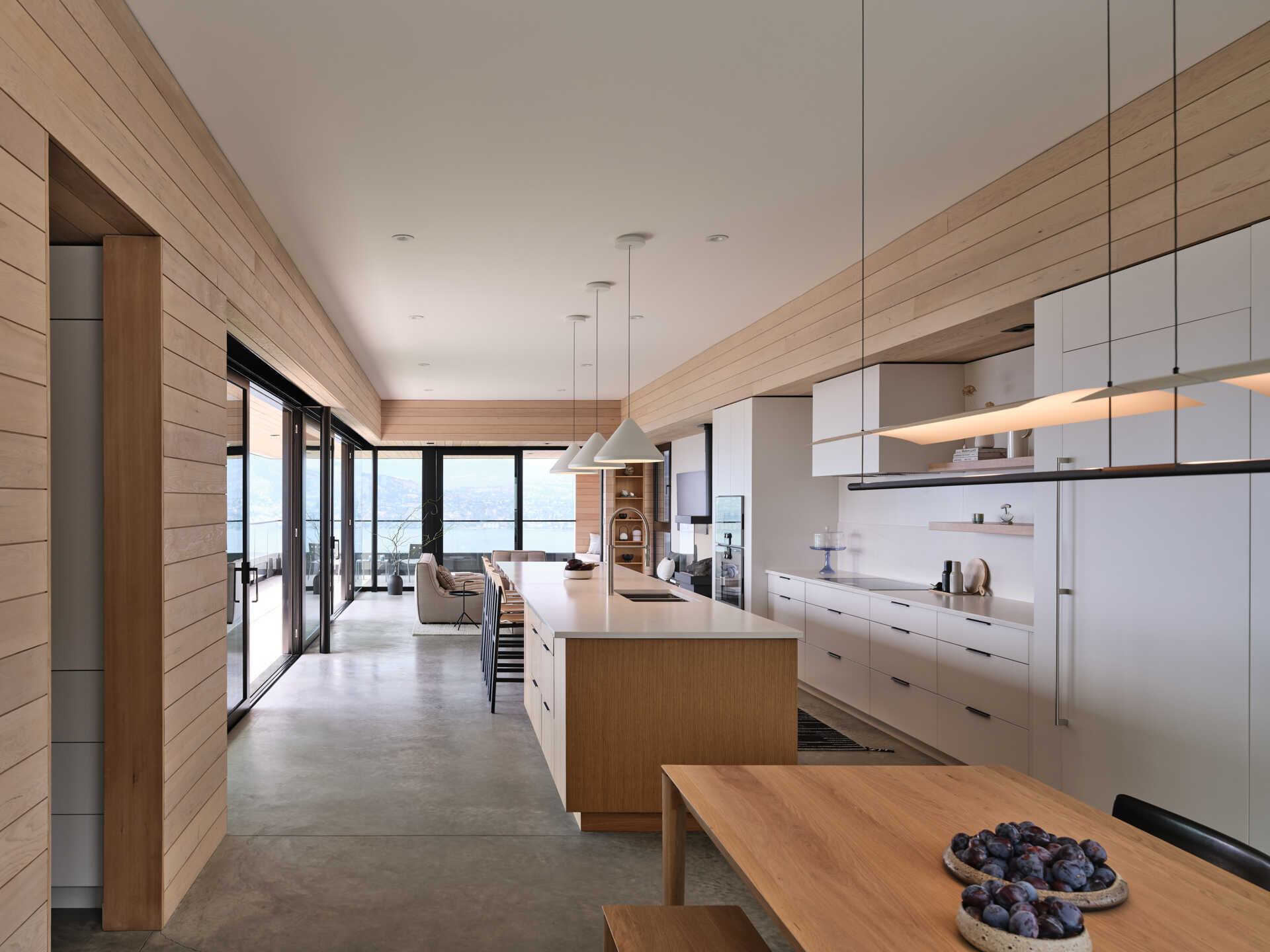 An open-plan modern kitchen, dining area, and living room arranged in one long space overlooking Okanagan Lake. White kitchen cabinetry, a wood island, warm wood wall paneling, and sliding glass doors define the interior.