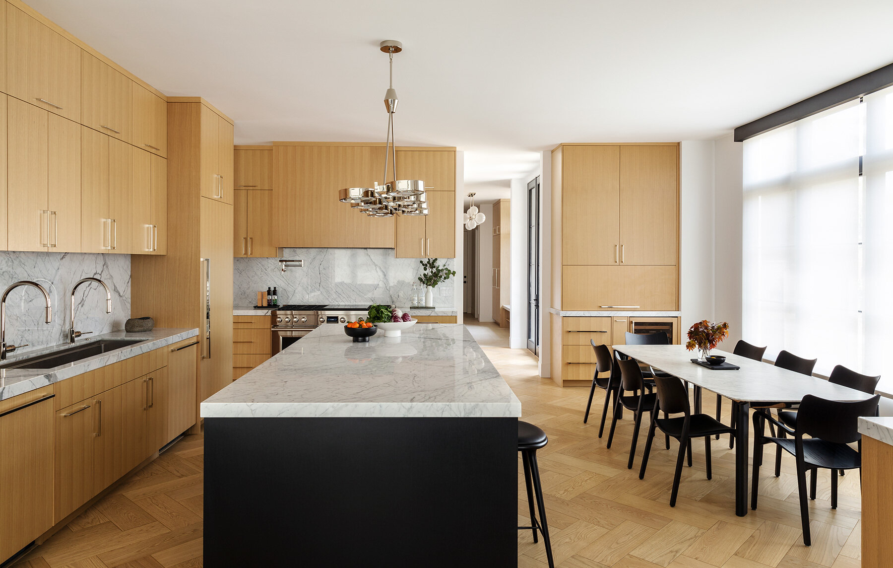 See the Atherton Residence kitchen and casual dining area by Paulina Carcach Studio, with pale timber cabinetry, veined stone surfaces, a black island, stainless-steel appliances and a relaxed dining table beside the windows.