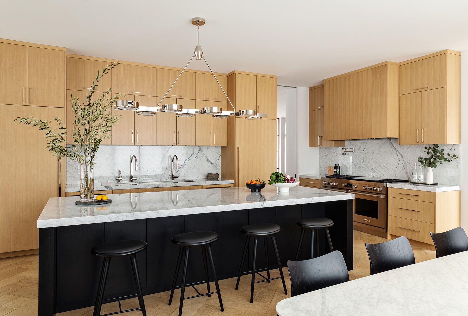 See the Atherton Residence kitchen and casual dining area by Paulina Carcach Studio, with pale timber cabinetry, veined stone surfaces, a black island, stainless-steel appliances and a relaxed dining table beside the windows.