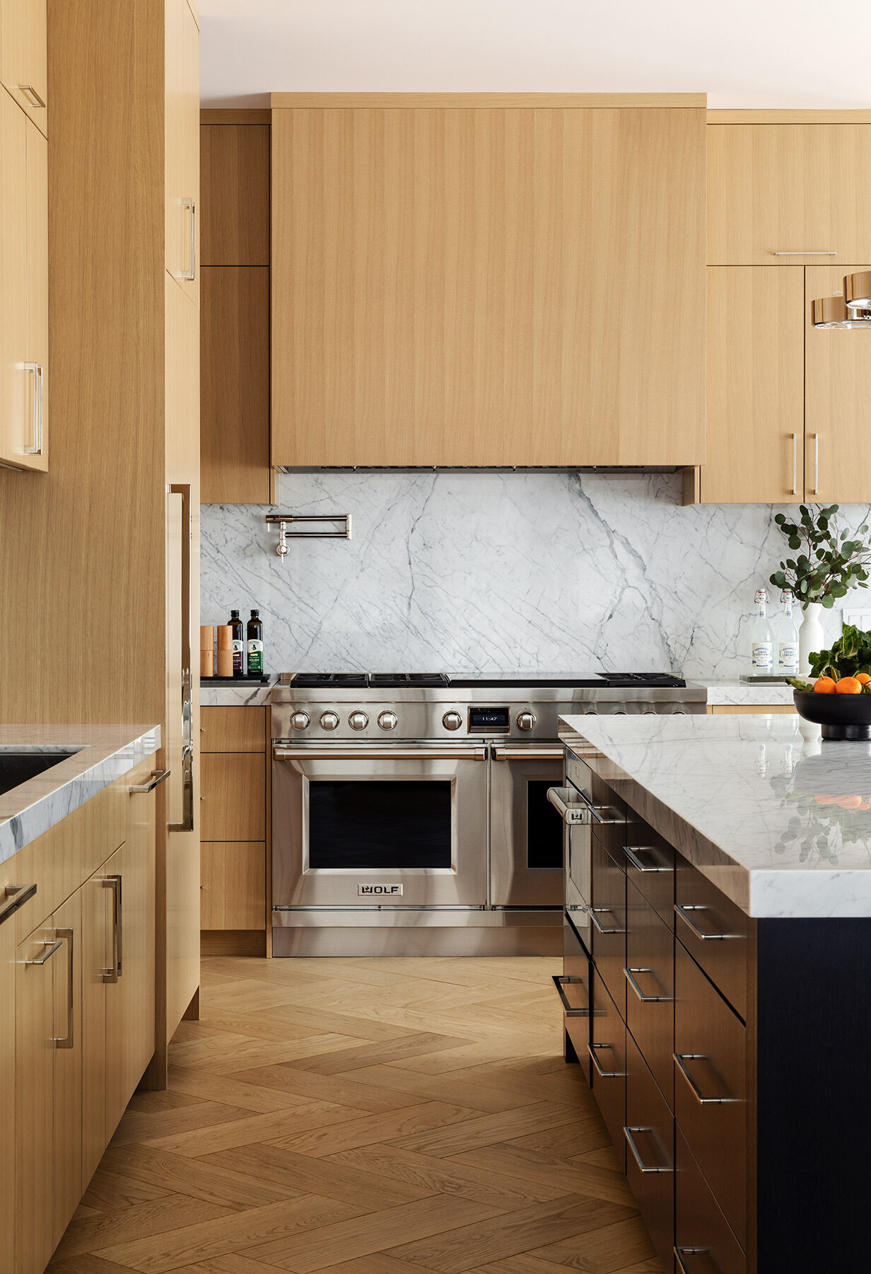 See the Atherton Residence kitchen and casual dining area by Paulina Carcach Studio, with pale timber cabinetry, veined stone surfaces, a black island, stainless-steel appliances and a relaxed dining table beside the windows.