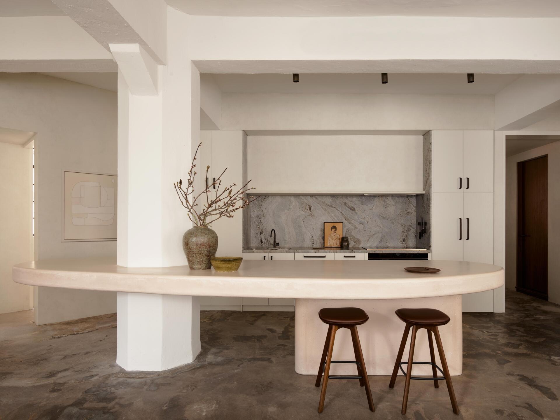A curved pale pink concrete kitchen counter wraps around an existing structural column in this Sydney apartment. Rounded edges, wood bar stools, white cabinets, and an industrial concrete floor create a modern kitchen with sculptural details.