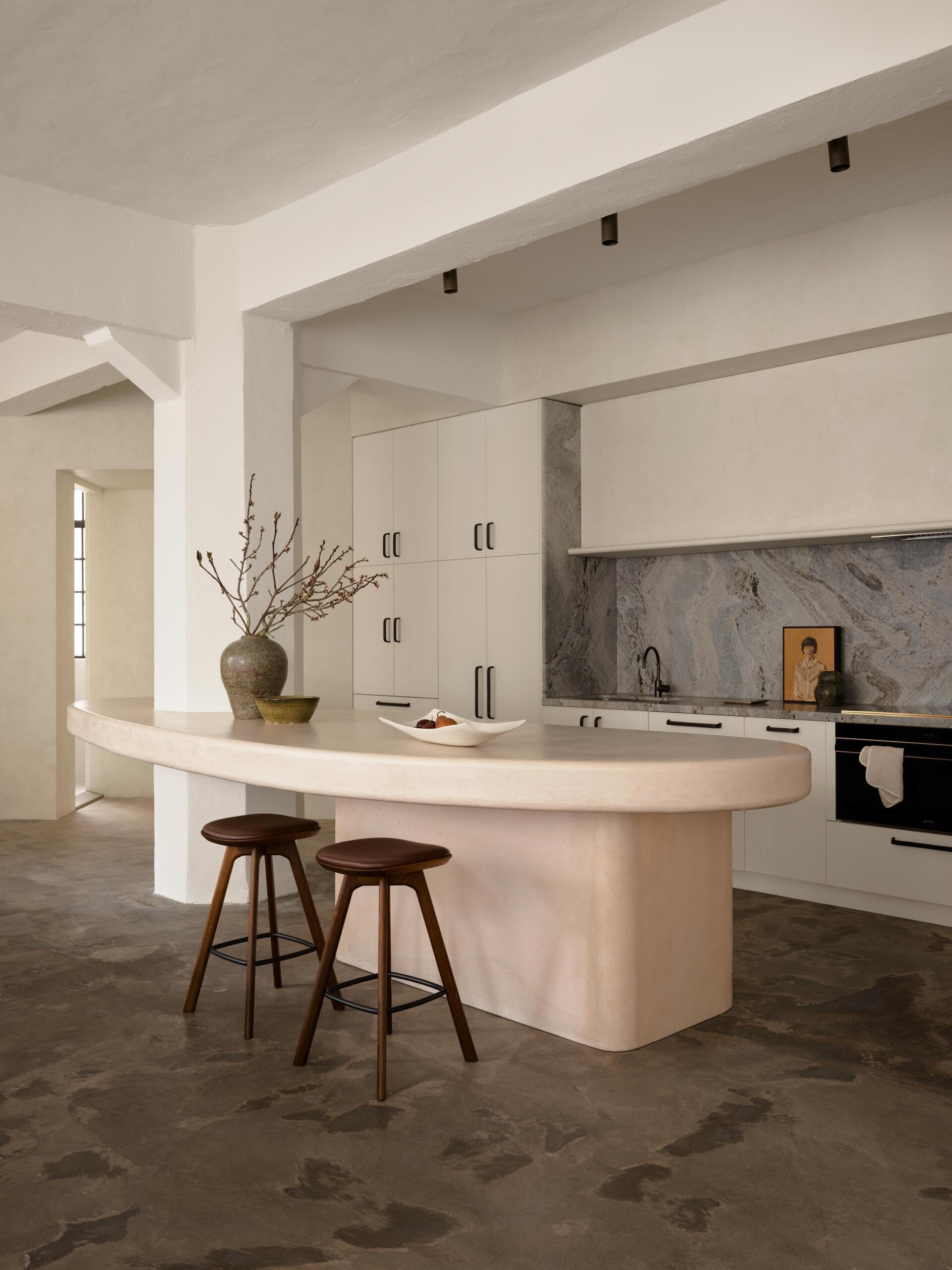 A sculptural kitchen counter with rounded edges and a pale pink concrete finish incorporates an existing column. Two dark wood bar stools, minimalist white kitchen cabinets, and a gray stone backsplash complete the contemporary apartment kitchen.
