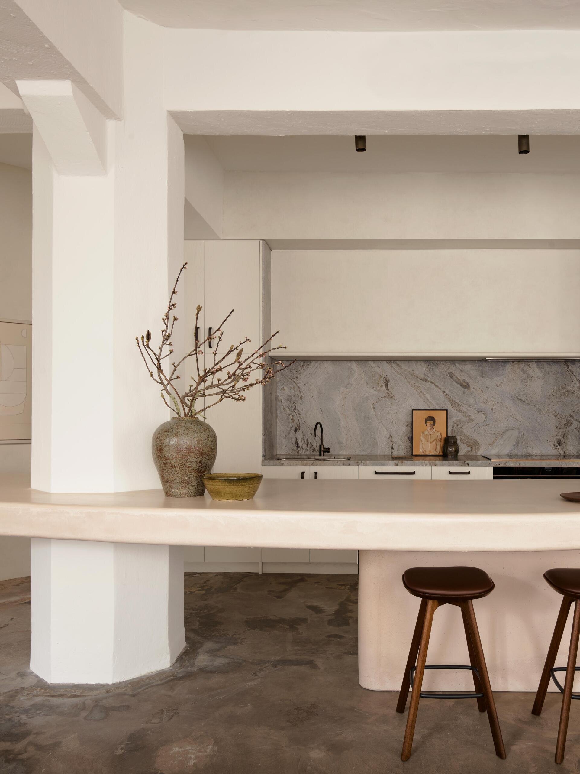 A sculptural kitchen counter with rounded edges and a pale pink concrete finish incorporates an existing column. Two dark wood bar stools, minimalist white kitchen cabinets, and a gray stone backsplash complete the contemporary apartment kitchen.