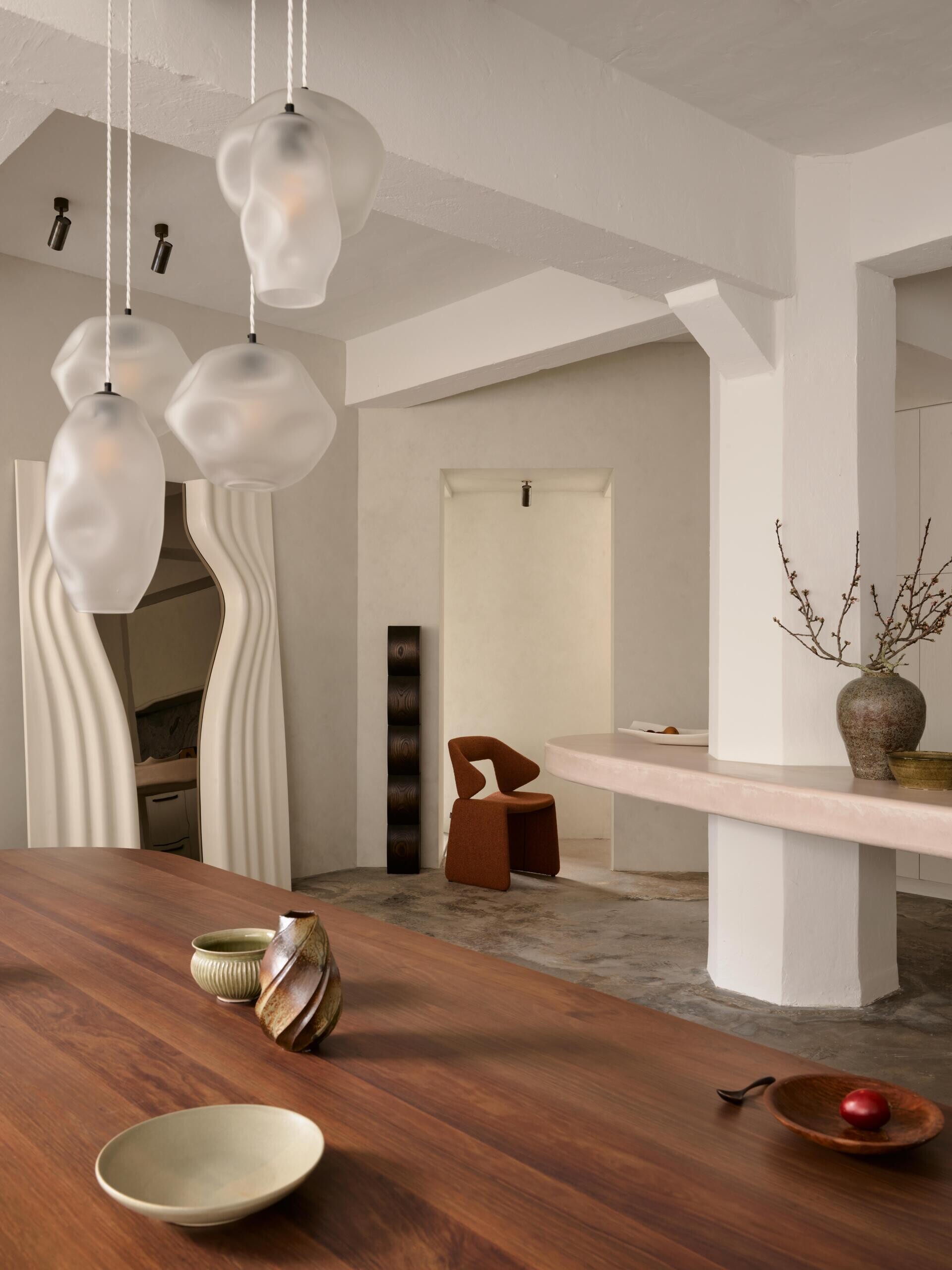 A modern open-plan dining and kitchen area featuring a curved concrete counter built around a white structural column. Sculptural pendant lights, a dark wood dining table, and a wavy white doorway add distinctive shapes to the industrial-inspired interior.