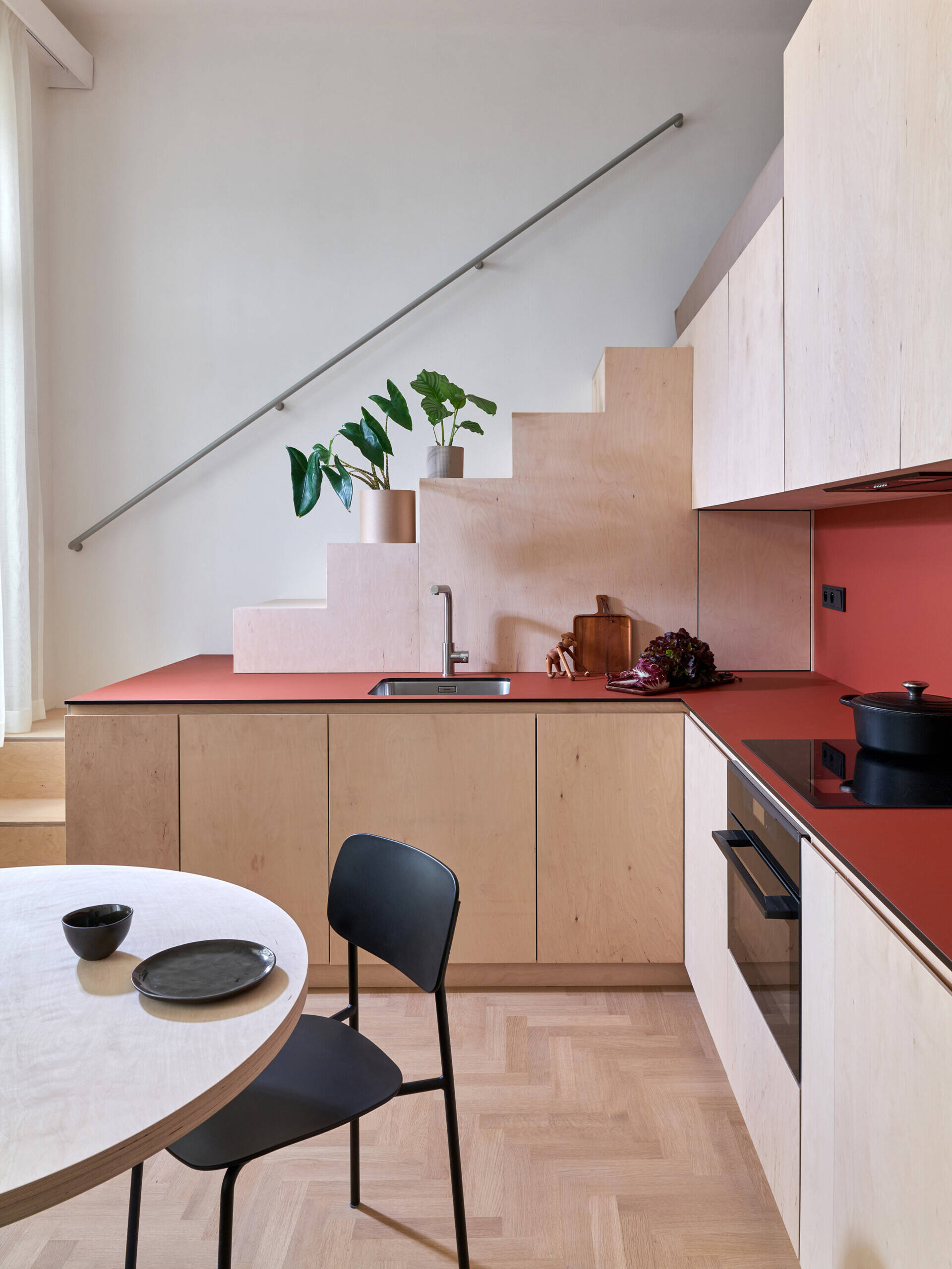 A compact apartment kitchen with pale cabinetry, red countertops, and a staircase integrated into the kitchen design. The steps lead to a mezzanine sleeping area above.