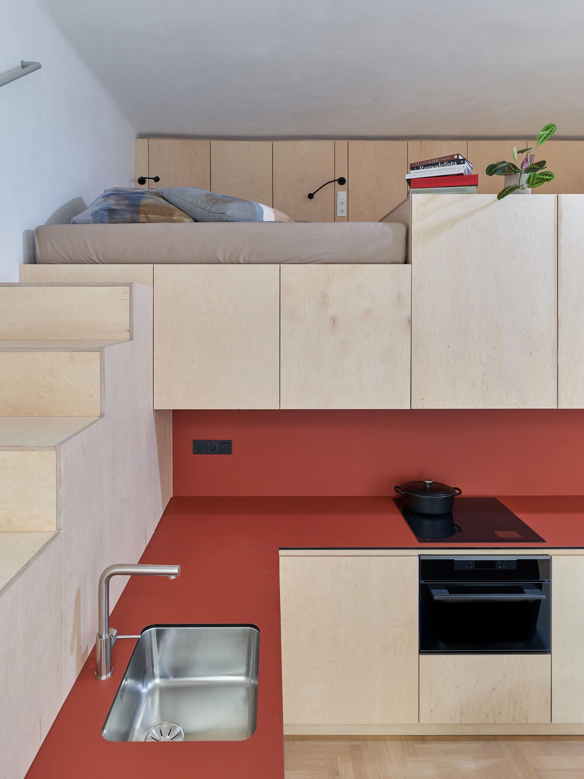 A loft bed positioned directly above a compact kitchen, with pale cabinetry, red countertops, and a built-in staircase. The vertical arrangement makes use of the studio apartment's limited floor space.