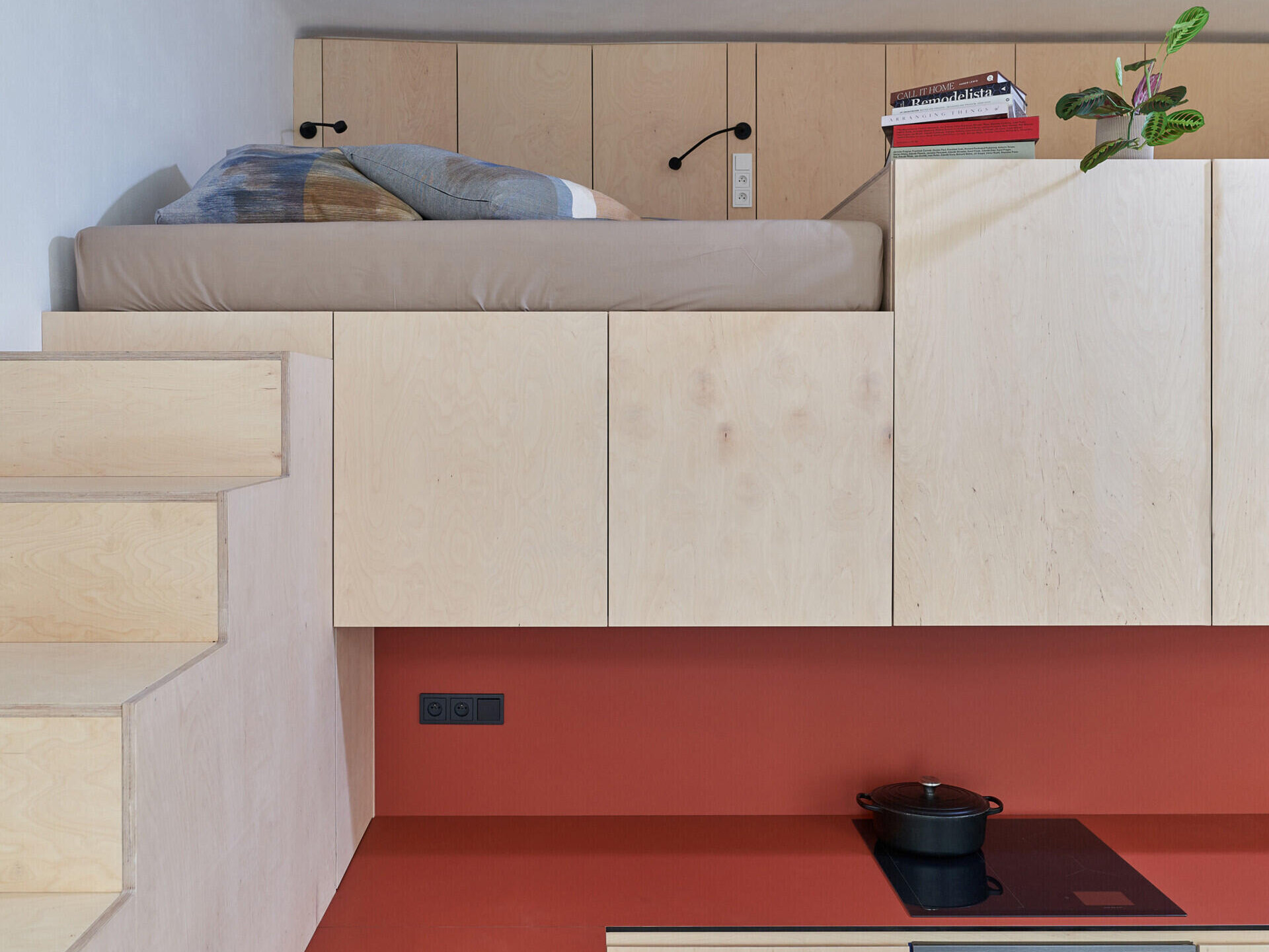 A compact studio apartment in Prague features a raised sleeping platform above a kitchen with red countertops and pale cabinetry. A built-in staircase connects the kitchen to the loft bed.