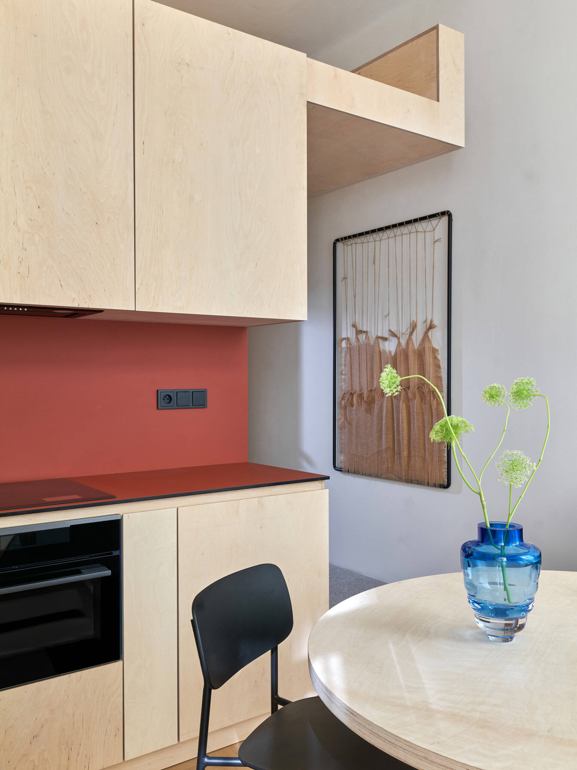 A small apartment dining area with a round table, black chair, pale kitchen cabinets, and a red backsplash. A raised sleeping platform extends above part of the kitchen.