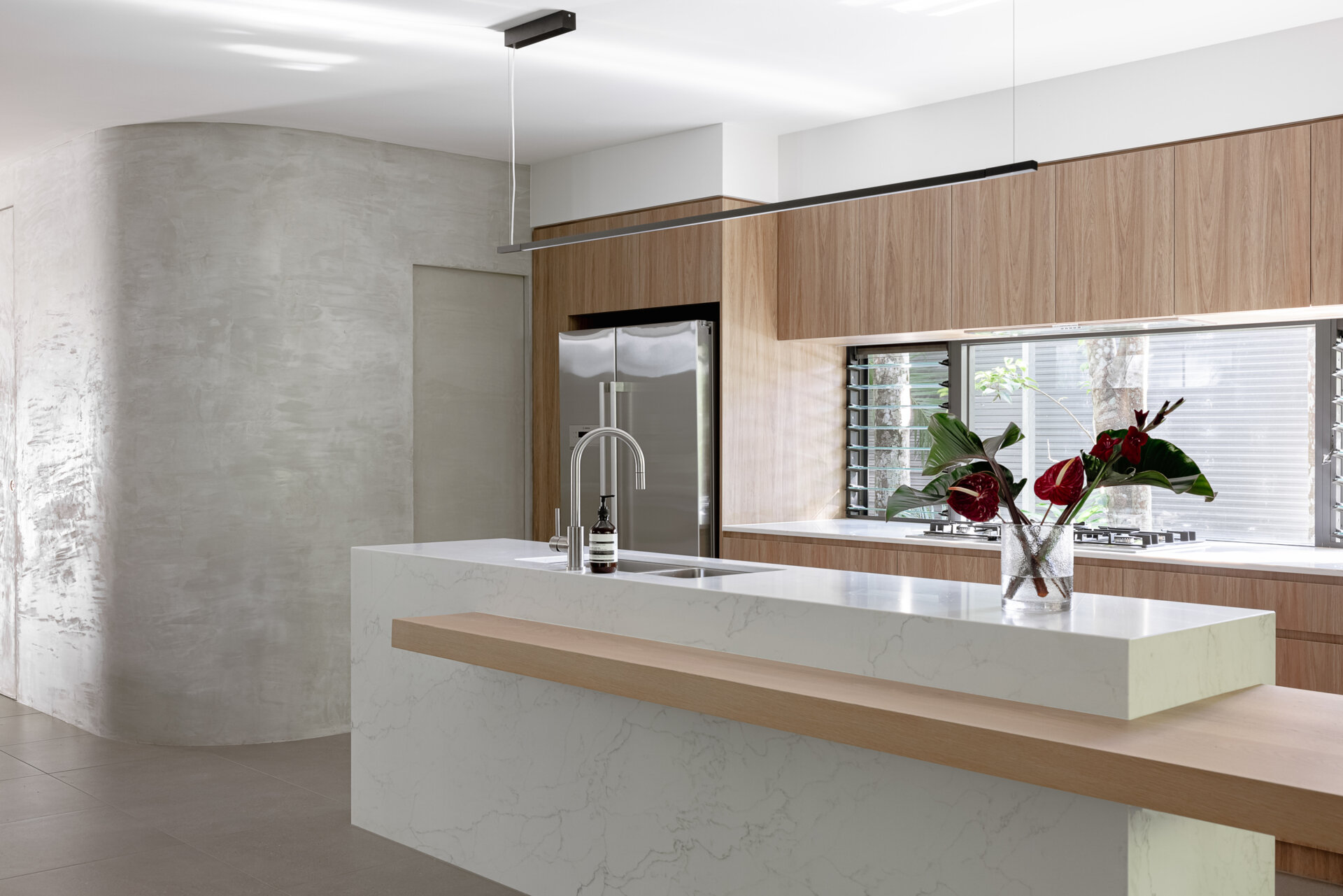 Explore the modern kitchen at Raintree in Noosa Heads, Australia, featuring natural timber cabinetry, a white marble-look island, an extended timber breakfast bar, integrated appliances, and a curved plaster wall.
