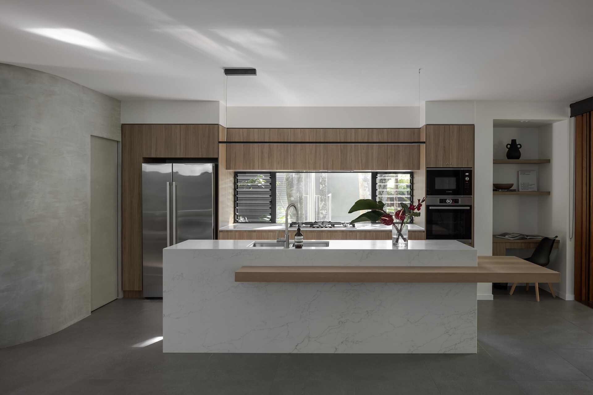 Explore the modern kitchen at Raintree in Noosa Heads, Australia, featuring natural timber cabinetry, a white marble-look island, an extended timber breakfast bar, integrated appliances, and a curved plaster wall.