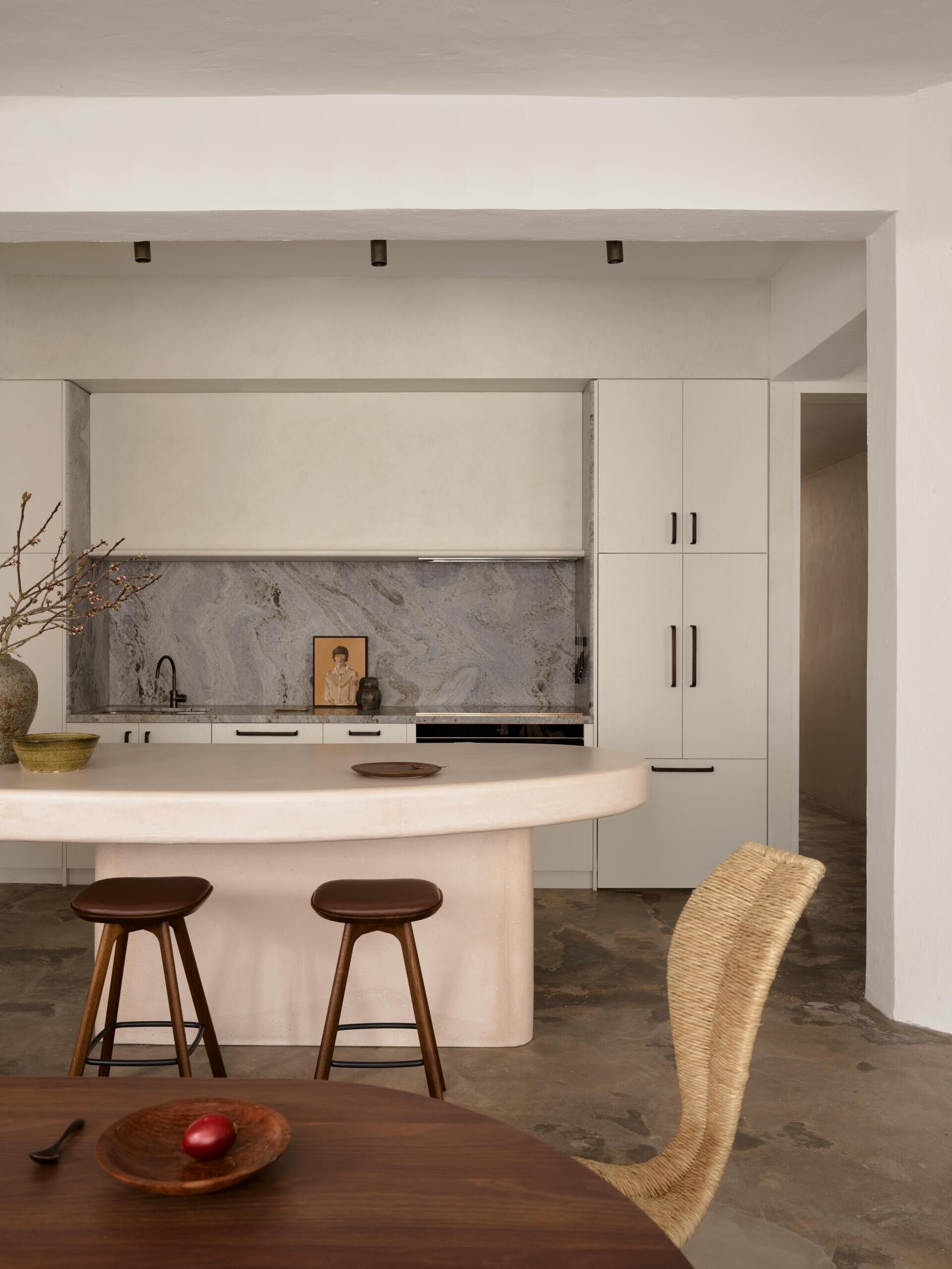 A contemporary kitchen with a long, pale concrete breakfast counter, rounded corners, white floor-to-ceiling cabinets, and a recessed gray stone backsplash. The original concrete floor and white structural beams preserve the apartment's industrial character.