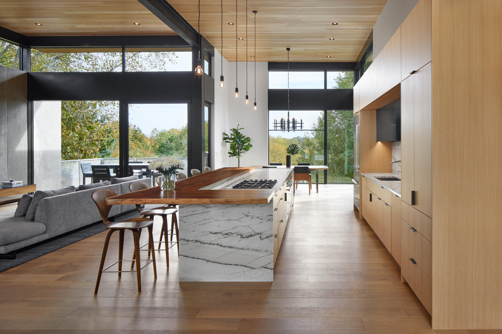 A contemporary kitchen featuring a long white stone island with gray veining, an extended wood breakfast bar, and light wood cabinetry. Floor-to-ceiling windows and high ceilings open the kitchen to the surrounding landscape.