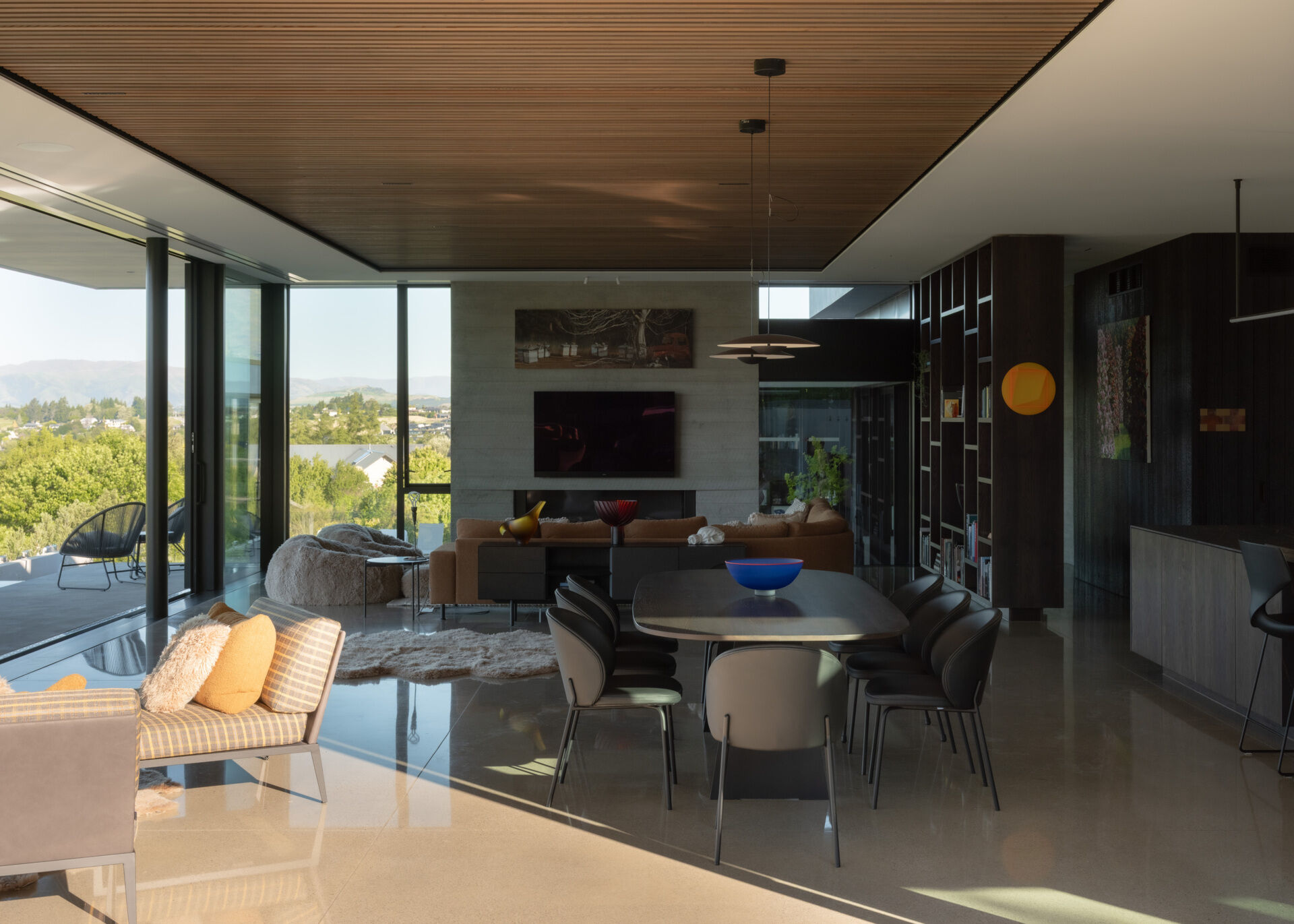 An open-plan living and dining room with polished concrete floors, a wood-slat ceiling, dark oak shelving, contemporary furniture, and colorful New Zealand artwork.